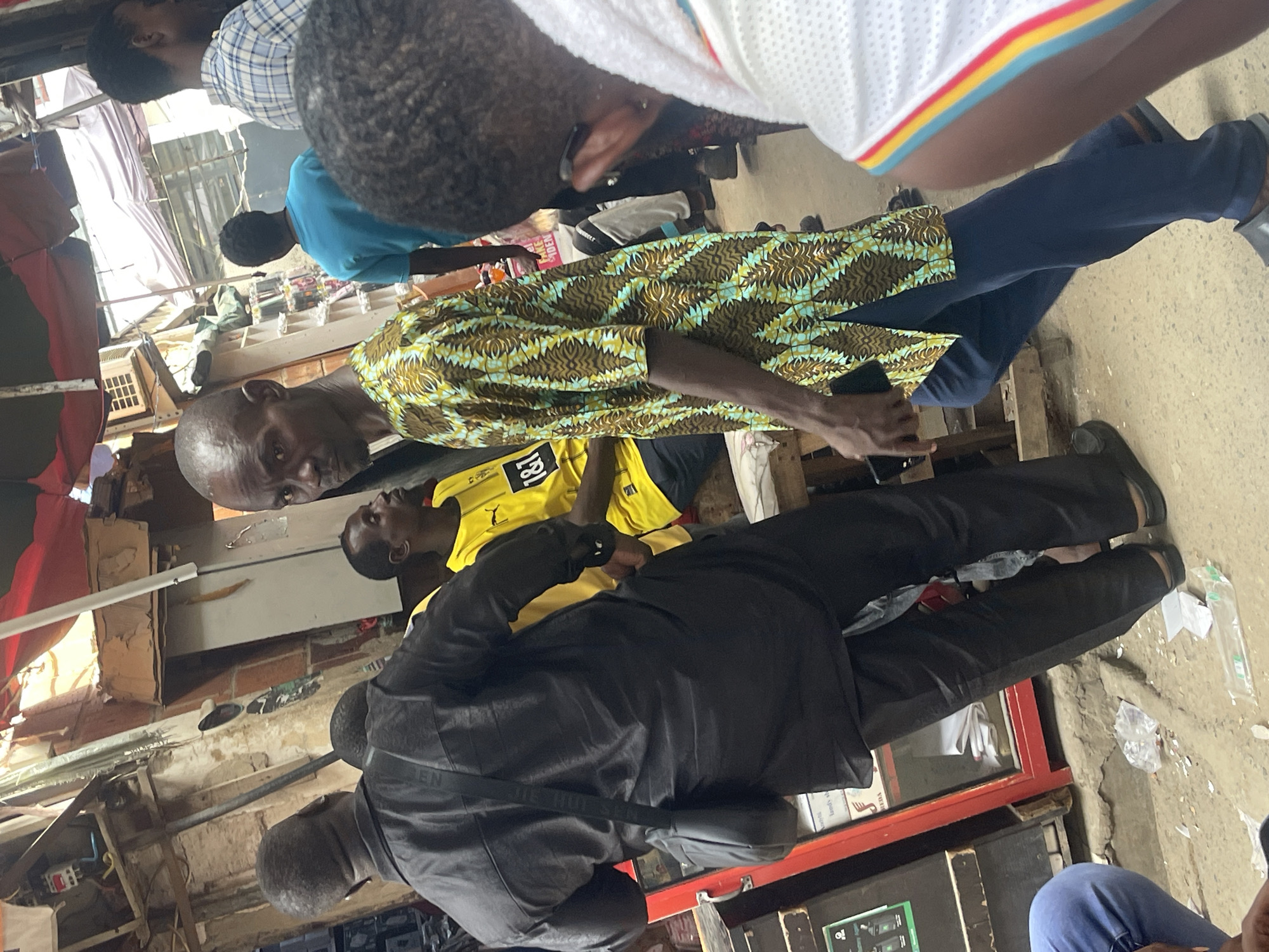 The image shows a group of people in a bustling outdoor environment, possibly a market scene in Abuja, Nigeria. One man is wearing a patterned green and brown shirt with blue pants, holding a phone. Another man is wearing a yellow soccer jersey with the number "181" on it, and someone in a black jacket is standing nearby. There are other people surrounding them. The background indicates a dense urban setting with shops and various structures.