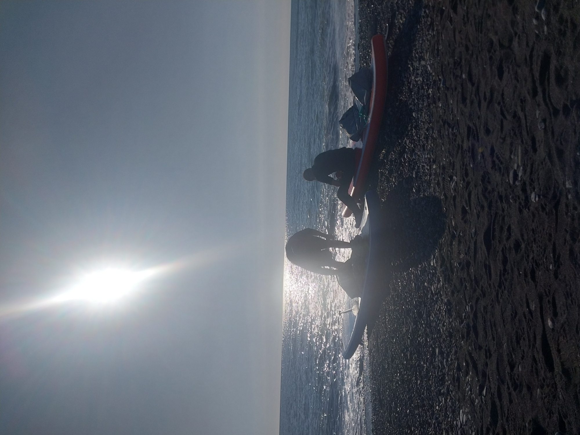 The image, oriented incorrectly, displays a bright coastal scene at either sunrise or sunset, indicated by the strong sun flare low on the horizon and the resulting backlighting. The sky is a hazy grey-blue on the left, brightening considerably where the sun shines.
On the right side of the image, a dark, pebbly or coarse-sand beach meets the ocean. Gentle waves lap at the shore, reflecting the intense sunlight with numerous sparkles.
Two individuals are visible, mostly silhouetted against the bright water and sky. One person is standing in the shallow water near a light-colored surfboard, appearing to be adjusting it or preparing to engage in a water sport. Further back, closer to the dark beach, another person is bending or kneeling near a prominent red-orange surfboard and what appears to be a bag or backpack. Both individuals are dressed in dark clothing, possibly wetsuits, suitable for water activities.
The scene suggests people preparing for or ending a surfing or paddleboarding session on a beach. No specific location cues or visible text are present. The atmosphere is bright and recreational.