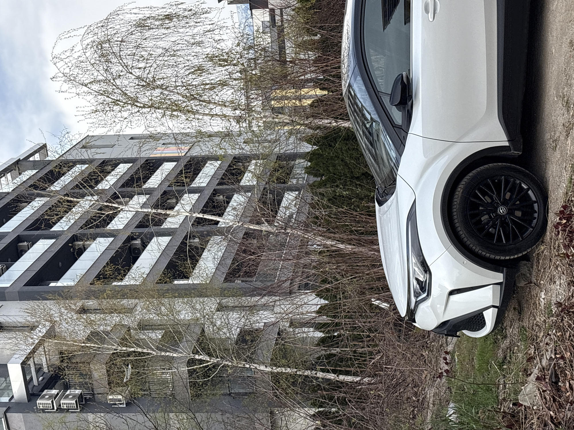 The image depicts a white car parked on a paved surface in an urban environment. The car's front passenger side is prominently featured, showcasing a black wheel. A modern building with multiple stories and reflective windows stands in the background. Bare trees with sparse foliage are scattered between the car and the building. The sky is overcast. Based on the architecture and vegetation, the location is likely in a residential or commercial area of Sofia, Bulgaria.