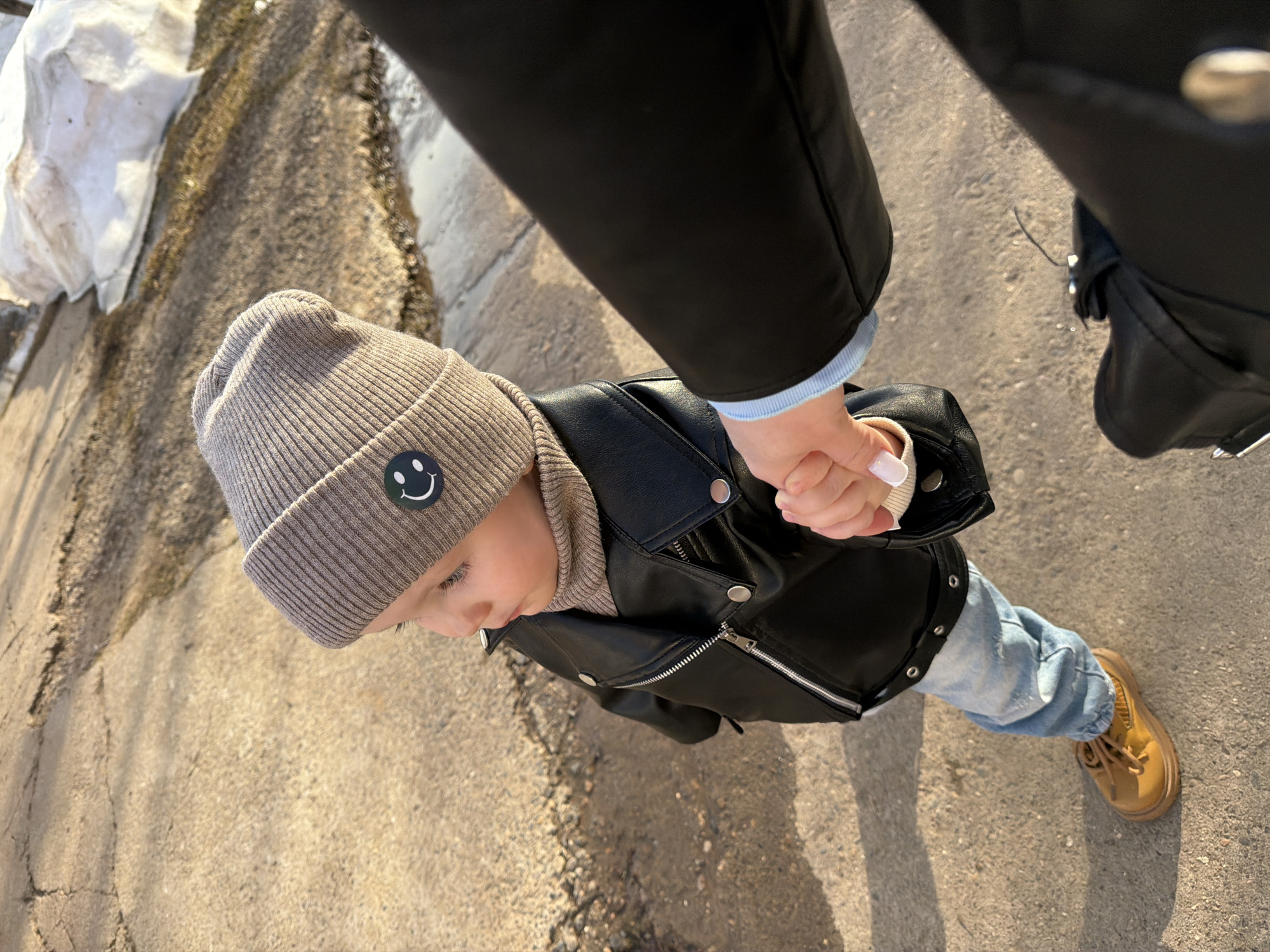 An overhead shot shows a child holding hands with an adult on a concrete surface. The child wears a grey beanie with a smiley face patch, a brown turtleneck, a black leather jacket, light blue jeans, and brown shoes. The adult wears a black jacket with a light blue cuff showing. The concrete surface has cracks and is partially shaded. A white structure is visible in the upper left corner. The photo was taken outdoors in the city of Chaikovsky, Russia.