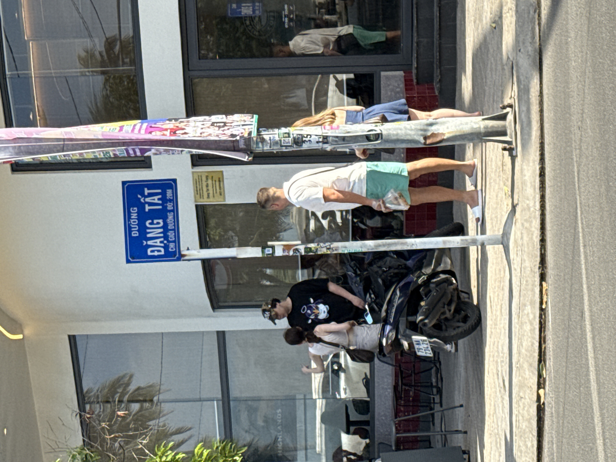 A street scene in Bac Nha Trang Ward, Vietnam, shows a sidewalk with a building in the background. A blue street sign with white lettering reads "ĐƯỜNG ĐẶNG TẮT CHỈ GIỚI ĐƯỜNG ĐỔ: 20M". A metal pole with various papers and stickers attached stands in the foreground.
Three people are visible on the sidewalk. In the middle ground, a man wearing a black t-shirt with a cartoon character and a baseball cap is talking to a woman in a white top and light-colored skirt. A dark-colored scooter is parked next to them. To the right, a man in a white t-shirt and teal shorts walks away from the camera, carrying a plastic bag. Beside him, a woman in a blue dress stands near the pole. In the background, through the building's windows, reflections of palm trees and people are visible. A man in a light-colored shirt and green shorts is partially seen inside the building. The ground is a concrete sidewalk next to a paved street. Sunlight casts shadows on the sidewalk.