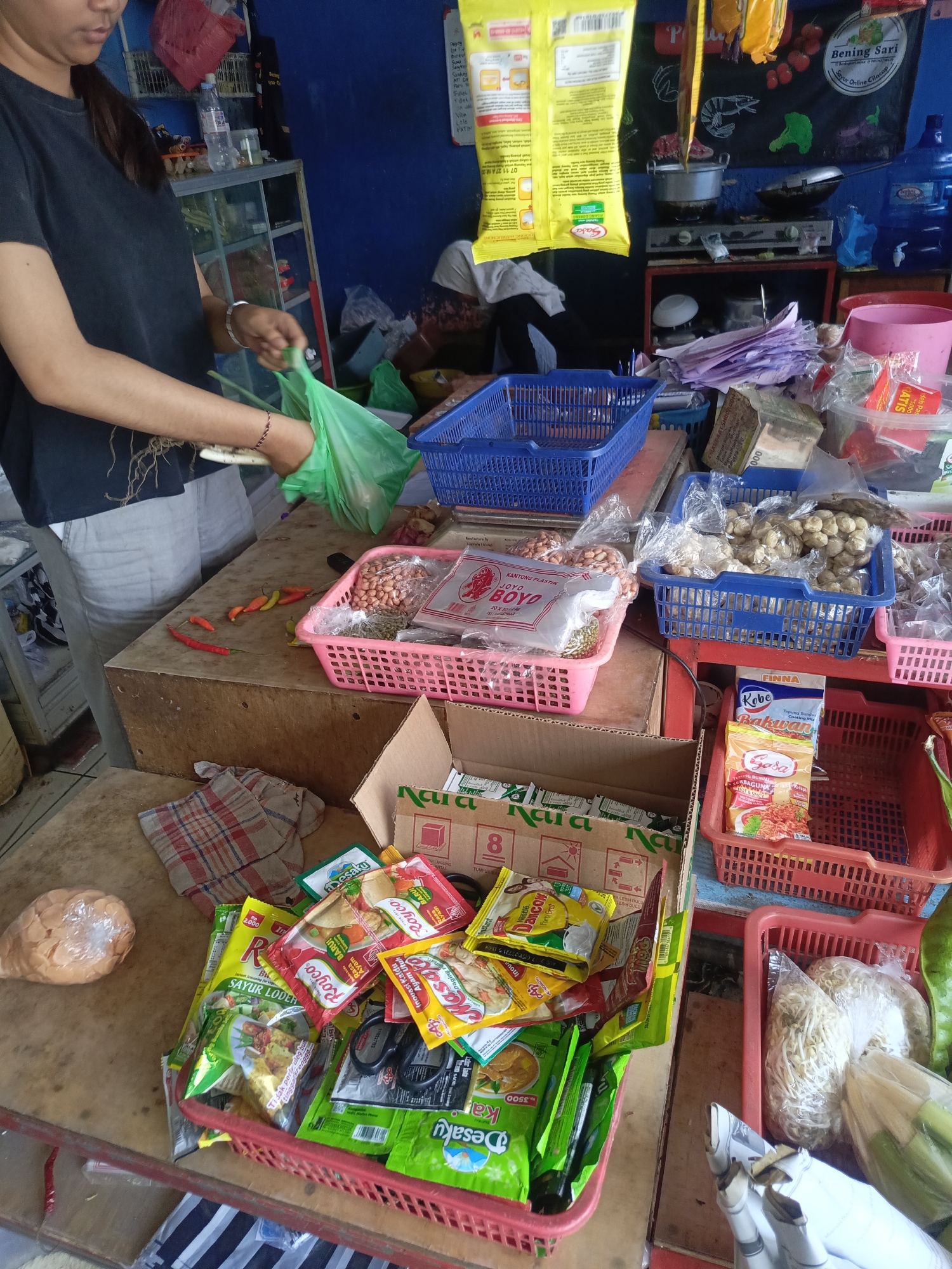 The image depicts an indoor scene, likely a small shop or market stall in Desa Gunungsimping, Indonesia. A person, partially visible from the chest up and wearing a dark t-shirt, is standing on the left side of the frame. They are holding a green plastic bag and appear to be in the process of bagging items. In front of them, on a wooden counter, are several red and yellow chilies. To the right of the chilies is a pink plastic basket filled with peanuts and other unidentifiable small items.
Behind the counter, various goods are displayed. There are several blue plastic baskets containing items like beans, spices, and other packaged foods. A gas stove is visible in the background, along with a large blue water jug. Yellow packaging for what appears to be instant noodles or seasoning mixes is hanging from above. A large red plastic container is also present, holding more packaged food items and a pair of scissors. Another pink plastic basket on the right side contains bean sprouts in plastic packaging.
The environment appears somewhat cluttered, with stacked boxes and items spread across the surfaces, suggesting a busy retail space. The lighting is ambient, with some shadows present.