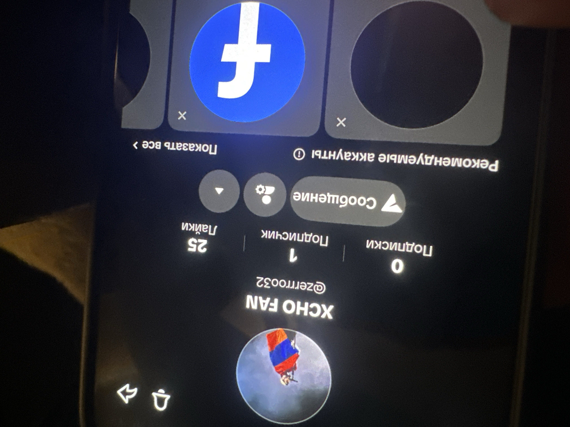 The image displays a smartphone screen showing a profile page on an app. The username is "XCHO FAN" with the handle "@zerrroo32" below it. The profile picture depicts a person skydiving with a parachute featuring red, blue, and orange colors.
Above the username, there are numerical statistics: 0 for "Podpiski" (Subscriptions), 1 for "Podpischik" (Followers), and 25 for "Layki" (Likes).
Above the statistics, there are three round icons: one with an arrow, another with a gear, and one with a play icon.
At the top of the screen, there's a section titled "Recommended accounts" (translated from Russian), featuring profile icons including a Facebook logo.
The overall aesthetic is dark, and the image quality appears to be captured in dim lighting.