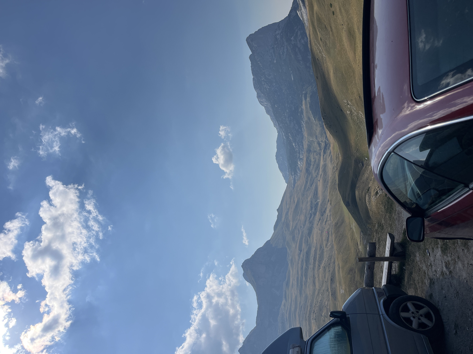 The image depicts a scenic mountain landscape under a clear blue sky with scattered clouds. In the foreground, two vehicles are parked. On the left is a silver van, and on the right is the back of a red car with its sunroof open. A wooden bench and post are visible between the vehicles, set against the backdrop of rolling hills and rugged mountains. The terrain appears arid and grassy, suggesting a high-altitude location. The overall scene conveys a sense of outdoor recreation or travel in a natural, mountainous environment. While the prompt mentions Dubrovsko, Montenegro, there are no specific visual cues in the image to confirm this exact location.