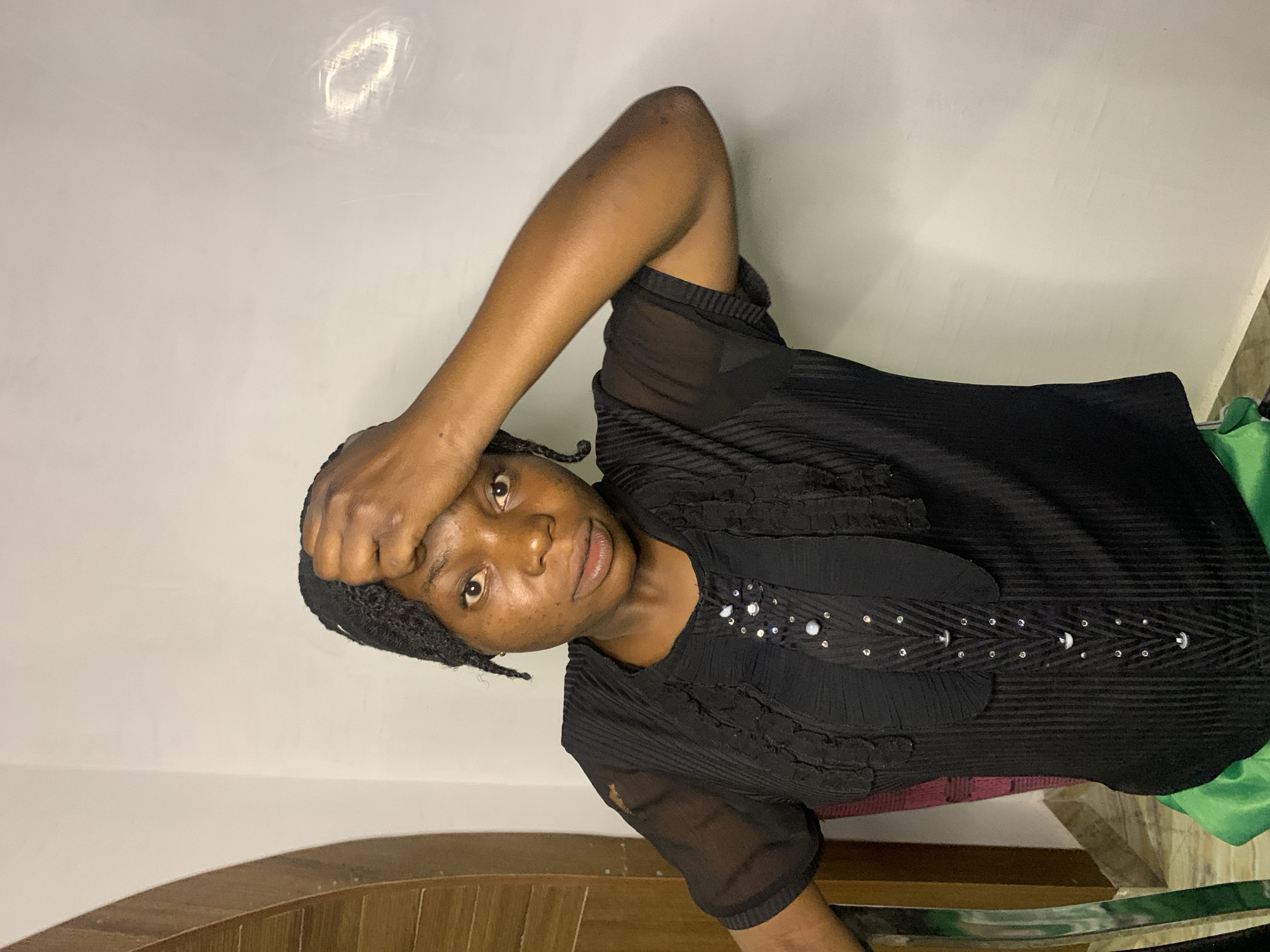 A young Black woman is seated indoors, her face turned towards the viewer. She is wearing a black, short-sleeved blouse with a ruffled neckline and decorative beadwork down the front. Her right hand is raised to her forehead, and her gaze is direct. The background consists of a textured white wall with subtle lighting, and to the left, a wooden architectural element and a sliver of what appears to be a chair with a maroon backrest are visible. A portion of bright green fabric is visible below her. The environment suggests an indoor setting, possibly a residence or studio. There are no specific indicators in the image to place the scene in the city of Jos, Nigeria.