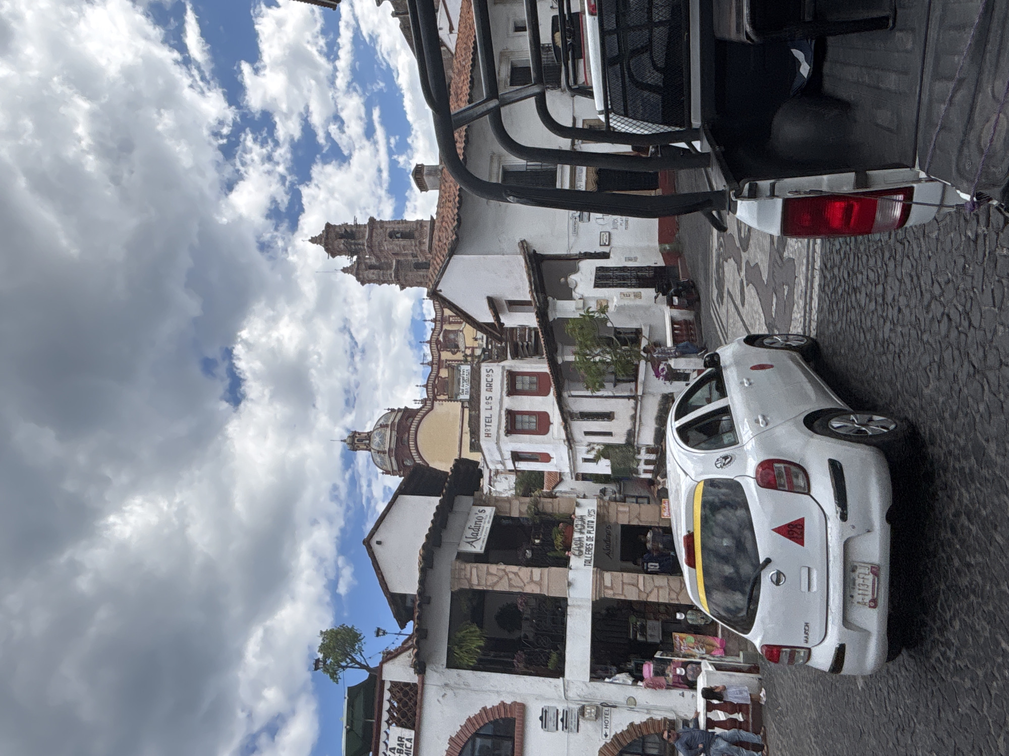 This is a photograph taken from a street-level perspective in Taxco, Mexico. The image is dominated by the architecture typical of the city, characterized by white buildings with red-tiled roofs and cobblestone streets. In the foreground, a white Nissan March taxi is parked on the left, its rear facing the camera. The taxi has a yellow stripe across its rear window and a red triangular decal with the number 420 on its trunk. Partially visible on the right is the rear of a white pickup truck, which appears to be a police vehicle due to the cage-like structure in its bed.
In the background, several buildings are visible, including one with a sign that reads "Aladino's" and "CASA ADJA TALLERES DE PLATA". Another building has a sign that says "HOTEL LOS ARCOS". Further in the distance, the iconic towers of the Santa Prisca Church can be seen against a cloudy sky. There are a few people walking on the street and sidewalks. The overall scene depicts a busy street in the historic center of Taxco, known for its silver shops and colonial architecture.