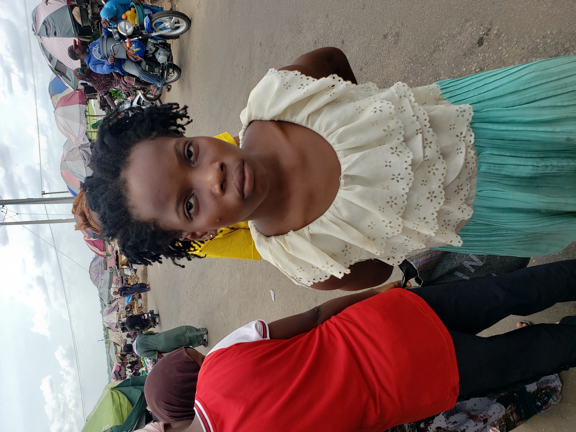 The image, rotated to vertical, depicts a lively outdoor market scene in Lokoja, Nigeria, under an overcast sky. In the foreground, a young Black woman is prominently featured, looking directly at the camera with a neutral expression. She wears a cream-colored, off-the-shoulder blouse with ruffles and intricate eyelet embroidery, paired with a light turquoise or mint green pleated skirt. Her dark, curly hair is visible.
Partially visible behind her to the lower right is another person wearing a red t-shirt with white trim and black trousers. The background reveals a busy market stretching along a paved road. Numerous market stalls are set up, each shaded by colorful umbrellas in various hues of blue, white, red, pink, and green. People are scattered throughout the market, some moving between stalls, others appearing to be vendors.
Two motorcycles are visible further into the market. One blue motorcycle is notably laden with baskets containing yellow and orange produce, possibly fruits. The cloudy sky suggests either early morning, late afternoon, or a generally overcast day, providing soft, diffused lighting. There is no clearly readable text visible within the image.