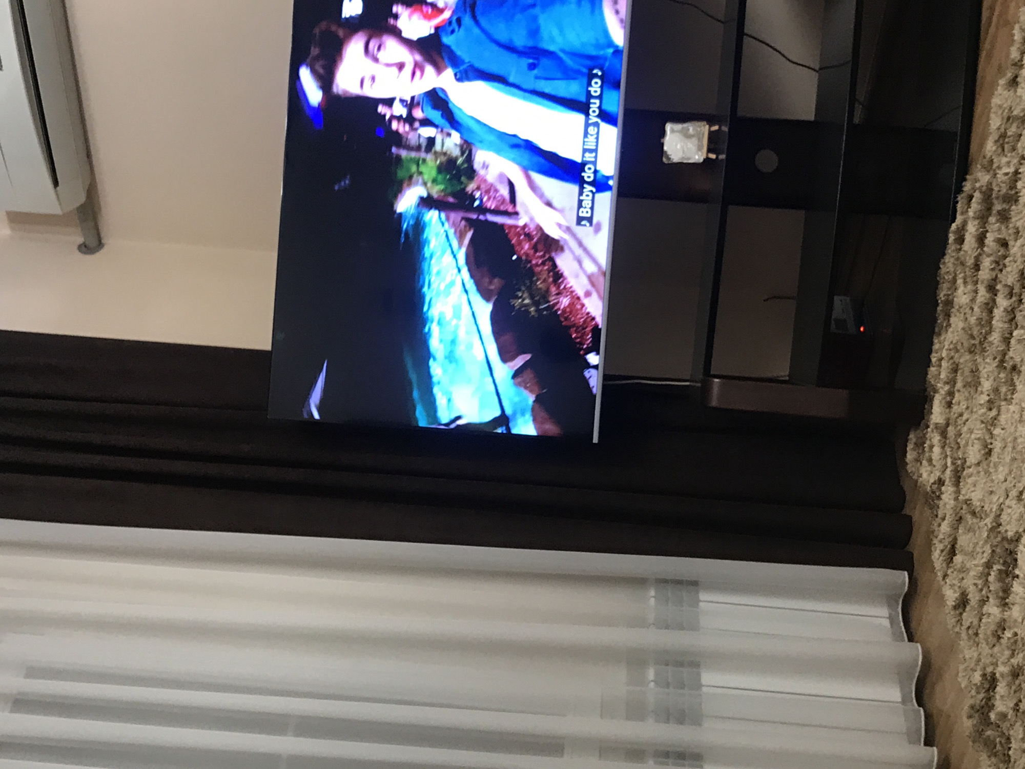 A horizontally oriented medium shot depicts a television displaying a musical performance, set within an interior space. The screen shows a fair-skinned individual in a blue jacket. The lyrics “Baby do it like you do do” are overlaid on the screen. The television sits on a dark-colored stand. The foreground includes a dark brown curtain, and white horizontal blinds. To the right, a section of a light-colored shag rug is visible. In the upper left corner, a white air conditioning unit is mounted on the wall. The location appears to be a domestic interior, potentially in Yerevan, Armenia.