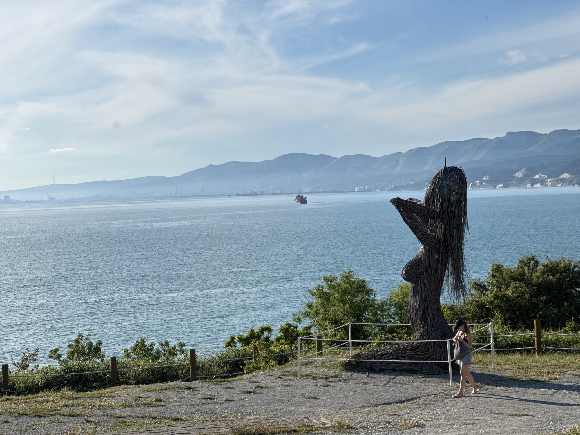 This panoramic image captures a sunny coastal landscape in Kabardinka, Russia, featuring a vast expanse of the sea, distant mountains, and a unique sculpture.
In the immediate foreground on the right, a large, dark sculpture resembling a reclining female figure is prominent. It appears to be constructed from interwoven natural materials like branches or reeds, with long, flowing elements suggesting hair. A woman, wearing a dark cap, a sleeveless top, and light-colored shorts, is seen walking past the sculpture, heading towards the left side of the frame. She carries a bag over her shoulder. A simple rope fence on wooden posts separates the gravelly viewing area from the lush green bushes and foliage on the slope leading down to the water.
The expansive, calm sea occupies the mid-ground, displaying gentle ripples on its blue-green surface. A solitary red cargo ship or tanker is visible sailing in the distance across the bay.
In the background, a long range of hazy blue mountains stretches across the horizon, their forms softened by atmospheric perspective. Along the base of these mountains, the distant coastline shows signs of urban and industrial development, including various buildings, cranes, and a distinct tall tower on the far left. The sky above is mostly clear with some wispy white clouds. The bright daylight suggests it is either morning or late afternoon. There is no visible text in the image.