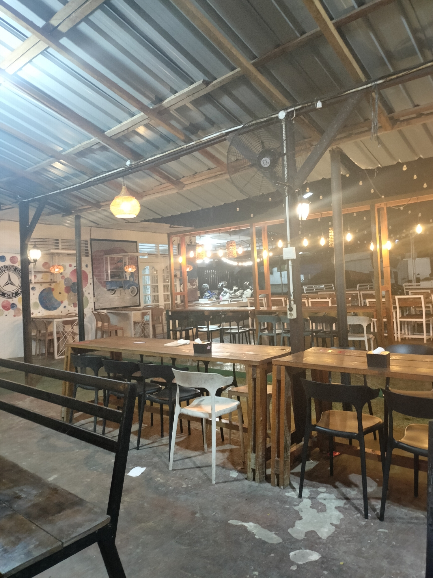 The image depicts an outdoor seating area of a cafe or restaurant, possibly located in Langsa, Indonesia. The scene is dimly lit, illuminated by hanging pendant lights and string lights that create a warm ambiance. The space is covered by a corrugated metal roof supported by wooden beams, suggesting an open-air or semi-outdoor setup.
Various tables and chairs are arranged throughout the area. In the foreground, a long wooden table with several dark chairs is visible. Further back, more tables and chairs, including some white plastic chairs, are present. The floor appears to be concrete, with some light-colored stains or puddles.
On the left side of the image, a wall features a circular logo with the text "Mercedes-Benz Club Aceh," and some colorful abstract art. There are also decorative pendant lights and what appears to be a large framed artwork depicting a retro blue vehicle.
In the background, through glass partitions, more seating areas and possibly a counter or kitchen area are visible. Several motorcycles are parked inside the establishment. A large industrial fan is mounted on a wooden support structure above the seating area. The overall impression is that of a casual dining establishment, possibly popular among motorcycle enthusiasts.