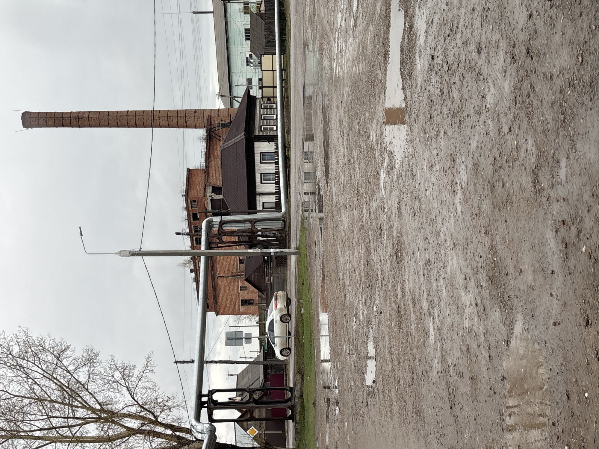 The image depicts a wet, muddy street scene in Ust-Ordynsky, Russia. A tall brick chimney stands prominently near several buildings, one with dark brown trim. A white car is parked near a grassy patch and a road sign. Overhead pipes cross the foreground. The sky is overcast, and a bare tree is visible in the lower left.