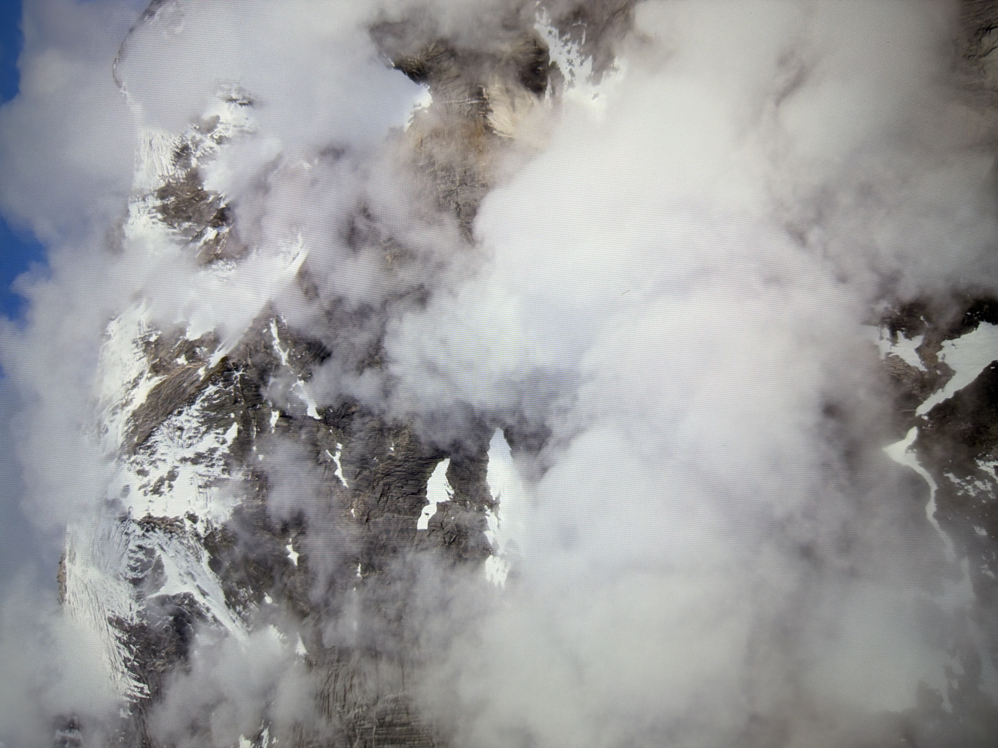 This is a photograph of a mountainous landscape partially obscured by clouds. Snow covers the peaks and slopes of the mountains. The sky is visible in the upper right corner, showing patches of blue. The image does not depict any urban environment, city, or specific location in Kyiv, Ukraine.
