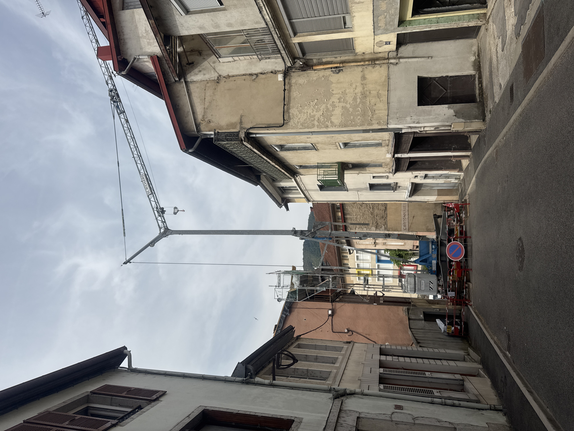 The image depicts a street in Saint-Claude, France, with buildings lining both sides and a construction crane dominating the upper portion of the scene. The buildings appear to be old, with weathered facades and varied architectural styles. The street is paved and appears to be in use, though some sections are cordoned off with red barriers, indicating ongoing construction or roadwork. A blue construction crane, identified by text as "DR. MAT" and "R.C. RICHARD", is positioned in the middle of the street, its arm extending across the buildings. Scaffolding is visible around a section of one building, suggesting renovation work. A blue crane with the text "DR. MAT" and "R.C. RICHARD" is present, suggesting construction activity. A traffic sign with a red circle and a horizontal blue bar, indicating no parking or stopping, is visible within the construction zone. The sky is overcast with clouds. The overall impression is one of urban life interspersed with construction and renovation.