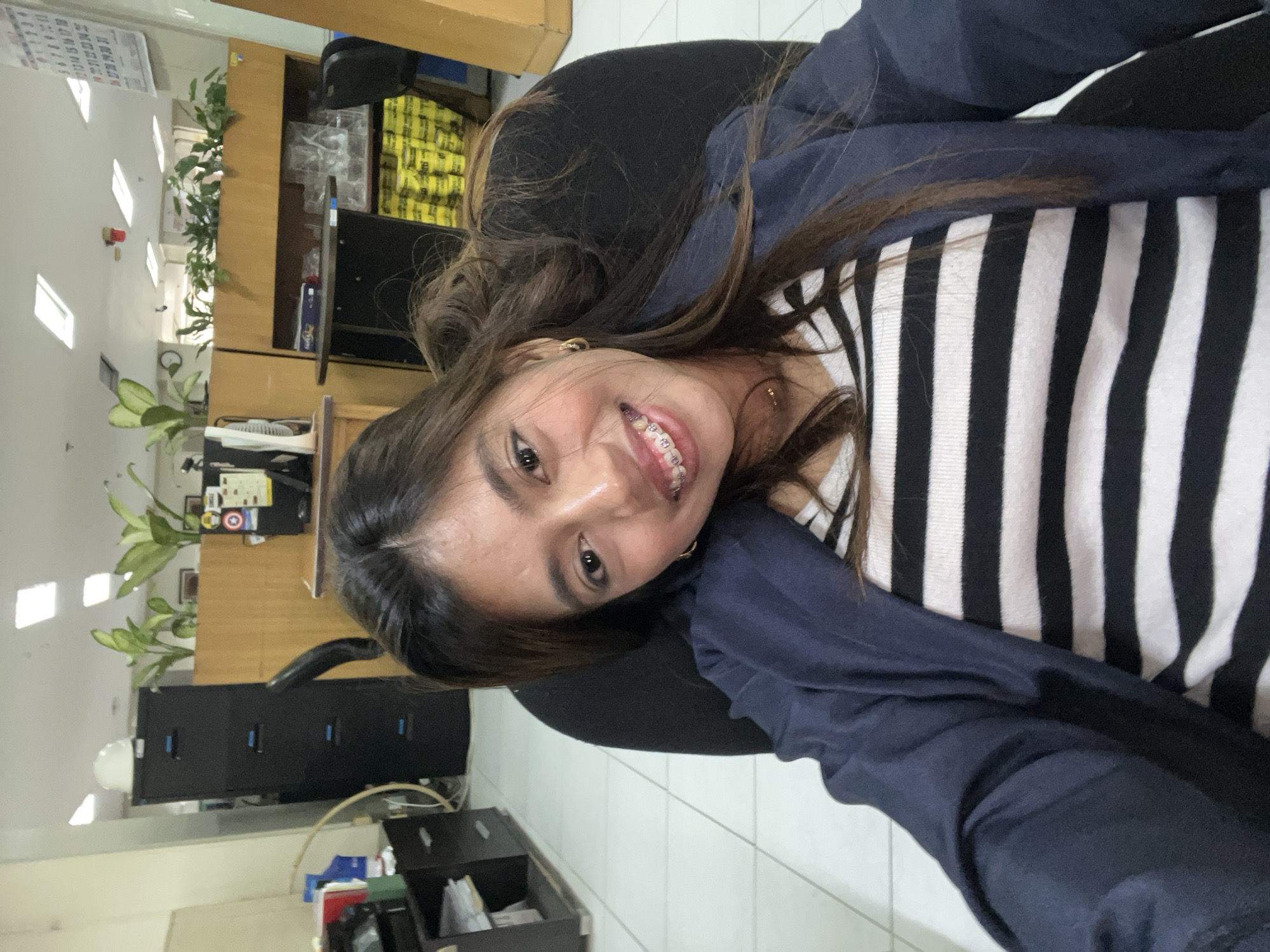 A young woman with long, dark hair is smiling at the camera. She is wearing a black and white striped shirt and a dark blue jacket. She has braces on her teeth and a gold necklace. She is sitting in a black office chair, and the background appears to be an office environment with filing cabinets, plants, and cubicle walls. The floor is tiled with white squares. The scene suggests a typical office setting, potentially in the city of Davao City, Philippines, given the context provided.