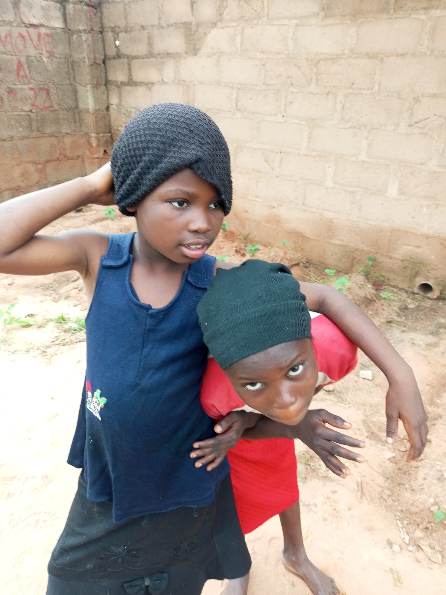 Two young girls, likely siblings, are pictured in what appears to be an outdoor, unpaved area adjacent to a building constructed of concrete blocks. The girl on the left, wearing a black knit cap and a dark blue sleeveless dress over a black skirt with a bow, is standing and has her right arm raised to touch her head. The girl on the right, wearing a red shirt and a black knit cap, is bent over and looking directly at the camera, with her arms around the other girl's waist. Both girls have dark skin and dark hair, and their faces are the focal point of the image. The background is a textured, light-colored brick wall with some red graffiti, and the ground is sandy with sparse vegetation. The scene suggests a casual, everyday moment in a residential or community setting. The implied location in Tempure, Nigeria, would place this scene in a West African urban or peri-urban environment.