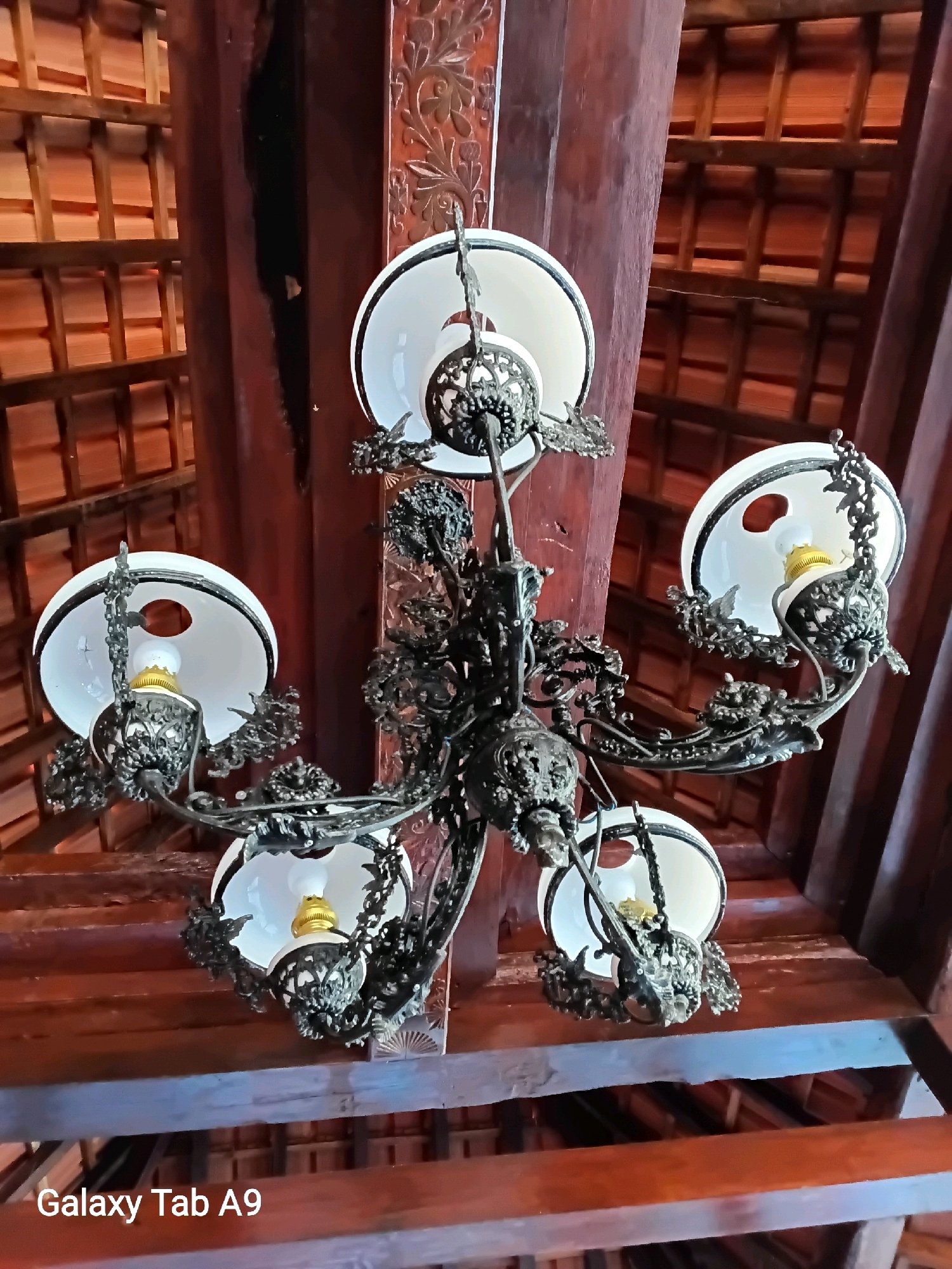 A vintage, ornate chandelier hangs from a wooden ceiling with visible beams and what appears to be exposed roofing tiles. The chandelier is made of dark metal with intricate scrollwork and features five white glass lampshades, each with a light bulb. The ceiling structure suggests a traditional or rustic architectural style. The image is a close-up, upward-facing shot of the chandelier and ceiling. The bottom left corner of the image displays the text "Galaxy Tab A9," indicating the device used for capture. The image was taken in Trenggalek, Indonesia.