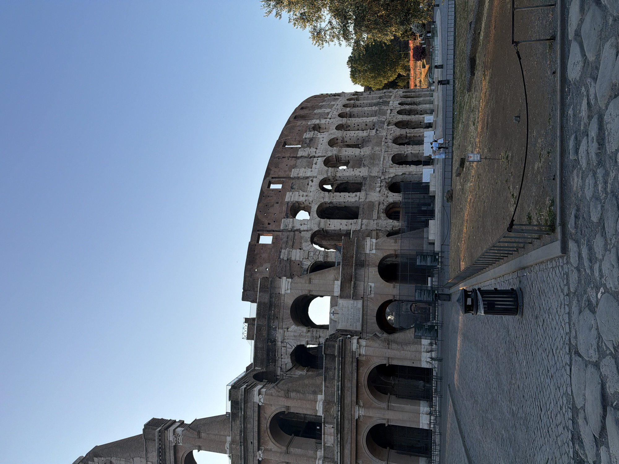 The image depicts the Colosseum in Rome, Italy, under a clear blue sky. The ancient amphitheater, a UNESCO World Heritage site, is a prominent feature, showcasing its weathered stone architecture with arched openings and sections of crumbling facade. In the foreground, a cobblestone pathway leads towards the Colosseum. Several metal fences delineate areas of the path and grassy sections. Two trash receptacles are visible on the path. In the midground, a small group of people, appearing to be tourists, stand near a fenced-off area. Trees are visible to the right of the Colosseum, bathed in natural sunlight. The scene suggests a daytime visit, with the lighting indicating either early morning or late afternoon.