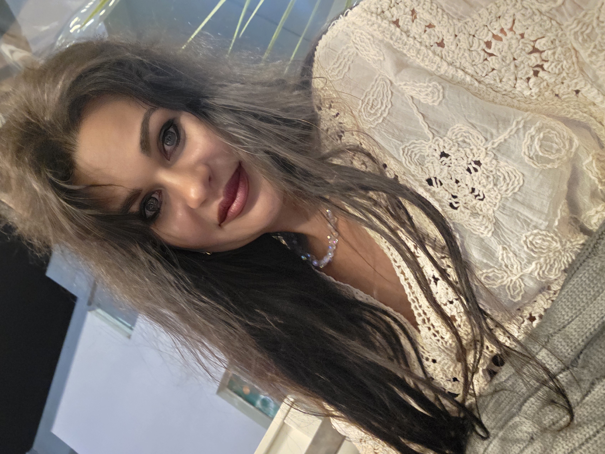 A woman with long, dark, and wavy hair is taking a selfie. She is wearing a white lace top and a pearl necklace. Her makeup is applied, with dark eyeliner and lipstick. In the background, there are some blurry elements that suggest an indoor setting with some greenery. The image does not contain any information about the city of Sochi, Russia.