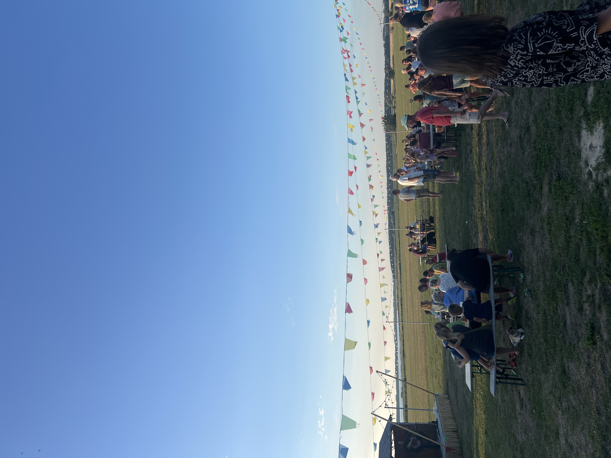 The image depicts an outdoor gathering at sunset near Am Salzhaff, Germany. A clear blue sky with sparse clouds forms the backdrop, with several strings of colorful pennant flags strung across the scene, some illuminated by string lights. In the foreground, the grass is mowed, with patches of dry soil.
To the left, a small stage structure is set up, with a person on it holding a guitar, suggesting live music. In front of the stage, a few people are seated at picnic tables. Further back, many people are scattered throughout the scene, standing and sitting around various picnic tables arranged on the grassy field. Some individuals appear to be eating or conversing. The middle ground shows a body of water, likely the Salzhaff, with a treeline visible on the horizon. The overall atmosphere suggests a relaxed, communal event, possibly a festival or a summer celebration.