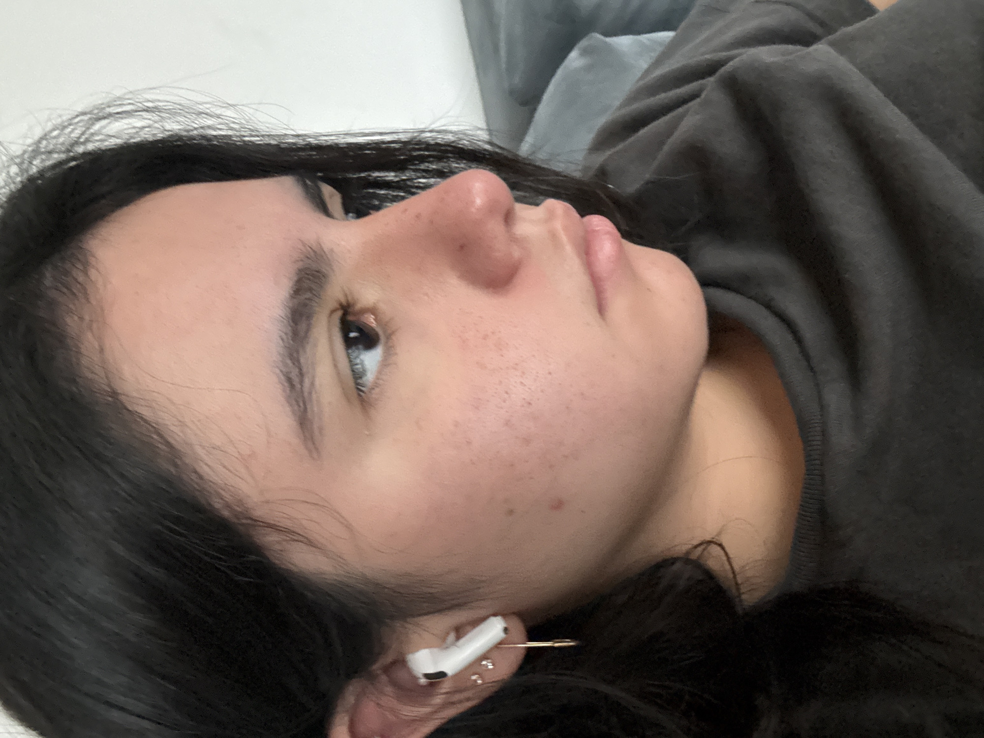 The image captures a close-up, side-profile portrait of a person, likely female, from the chest up. The person is lying down or resting, looking upwards and to the left with a neutral, relaxed expression.
**Subjects:** The central subject is a person with dark, wavy hair, fair skin, light-colored eyes (appearing blue, grey, or hazel), and full lips. Their face shows a scattering of small freckles or moles across the cheek and nose.
**Objects:** A white Apple AirPod is visible in the person's right ear, notably secured by a gold-toned safety pin piercing through the earlobe. Several small, shiny stud earrings are also visible in the earlobe. The person is wearing a dark grey or charcoal-colored long-sleeved garment, possibly a hoodie or sweatshirt, with visible ribbing detail around the neckline.
**Scene and Setting:** The scene is an indoor, close-up shot. The background on the upper left consists of a plain, light-colored surface, while the area behind the person's head and shoulder on the right is dark grey fabric, likely a pillow or blanket, suggesting a comfortable, relaxed setting.
**Activity or Event:** The person appears to be in a state of rest or contemplation, with their gaze directed upwards. The image seems to be either a candid moment or a posed shot emphasizing a relaxed posture.
**Notable Details:** The unique use of a safety pin to secure the AirPod is a prominent detail. The lighting is soft and even, typical of indoor conditions, making the time of day indeterminate. There are no specific outdoor weather cues or distinct location markers beyond the indoor setting. No visible text appears in the image.