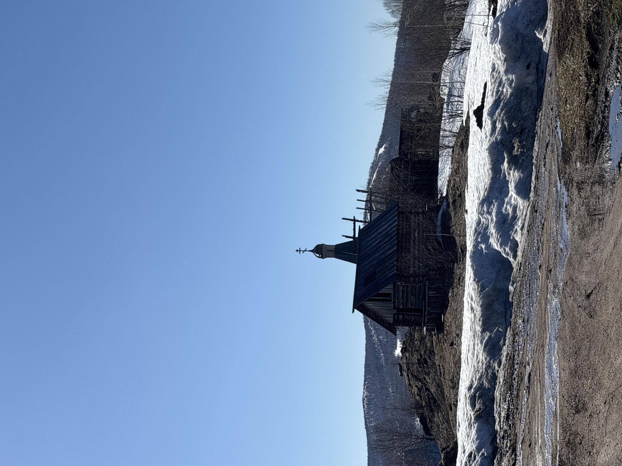 The image shows a wooden church standing near a snow-covered slope under a clear blue sky. The church has a distinct steeple with a cross on top. Part of a dirt road runs alongside the snow. The location is the Gubakha region in Russia.