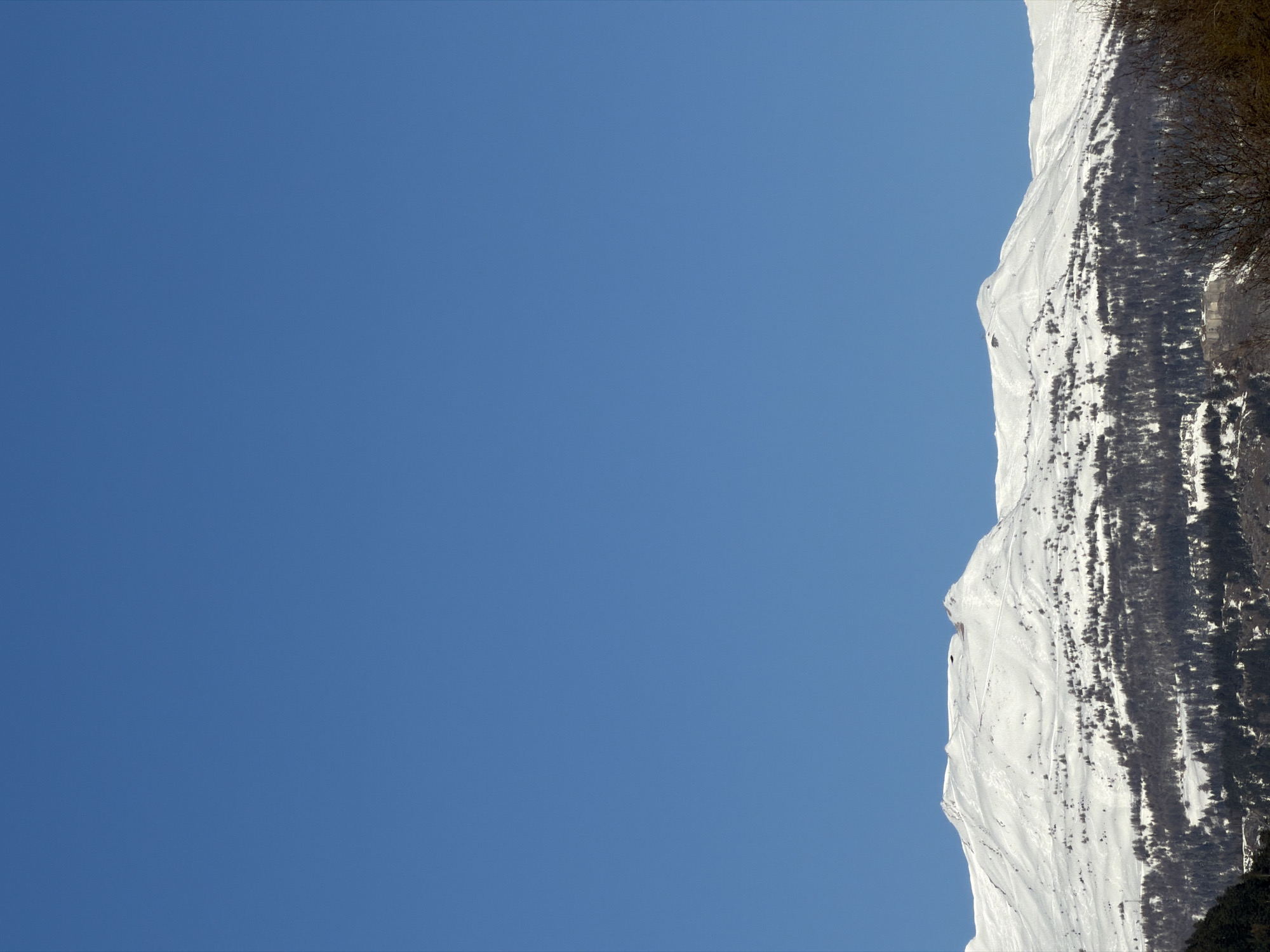 A landscape photograph shows a snow-covered mountain against a clear blue sky. The mountain occupies the right side of the frame, with the snowy surface extending down and slightly out of view at the bottom. Sparse vegetation and darker rock formations break up the white snow cover. The bright blue sky dominates the left side of the image. The location is possibly the French ski resort of Orelle.