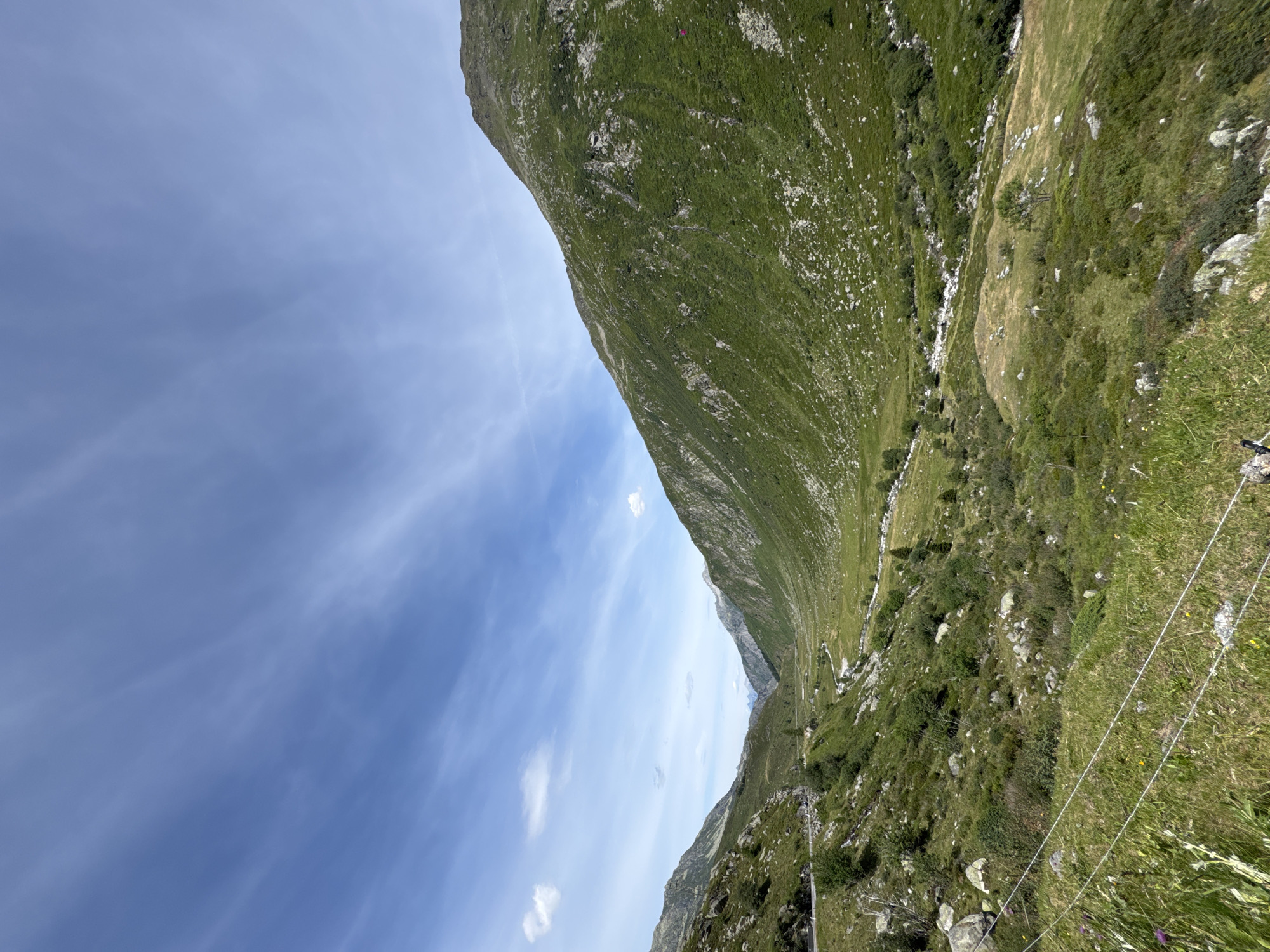 This is a daytime, eye-level, wide-angle photograph of a lush green mountain valley in Hospental, Switzerland. The sky is a clear blue with wispy white clouds. The valley is flanked by steep, grassy mountainsides dotted with rocks and sparse trees. A winding road is visible on the left side of the valley, and a small stream or river meanders through the valley floor. In the foreground, a wire fence runs horizontally across the image. There are no people or animals immediately visible in the scene. The overall impression is one of natural beauty and rugged terrain.