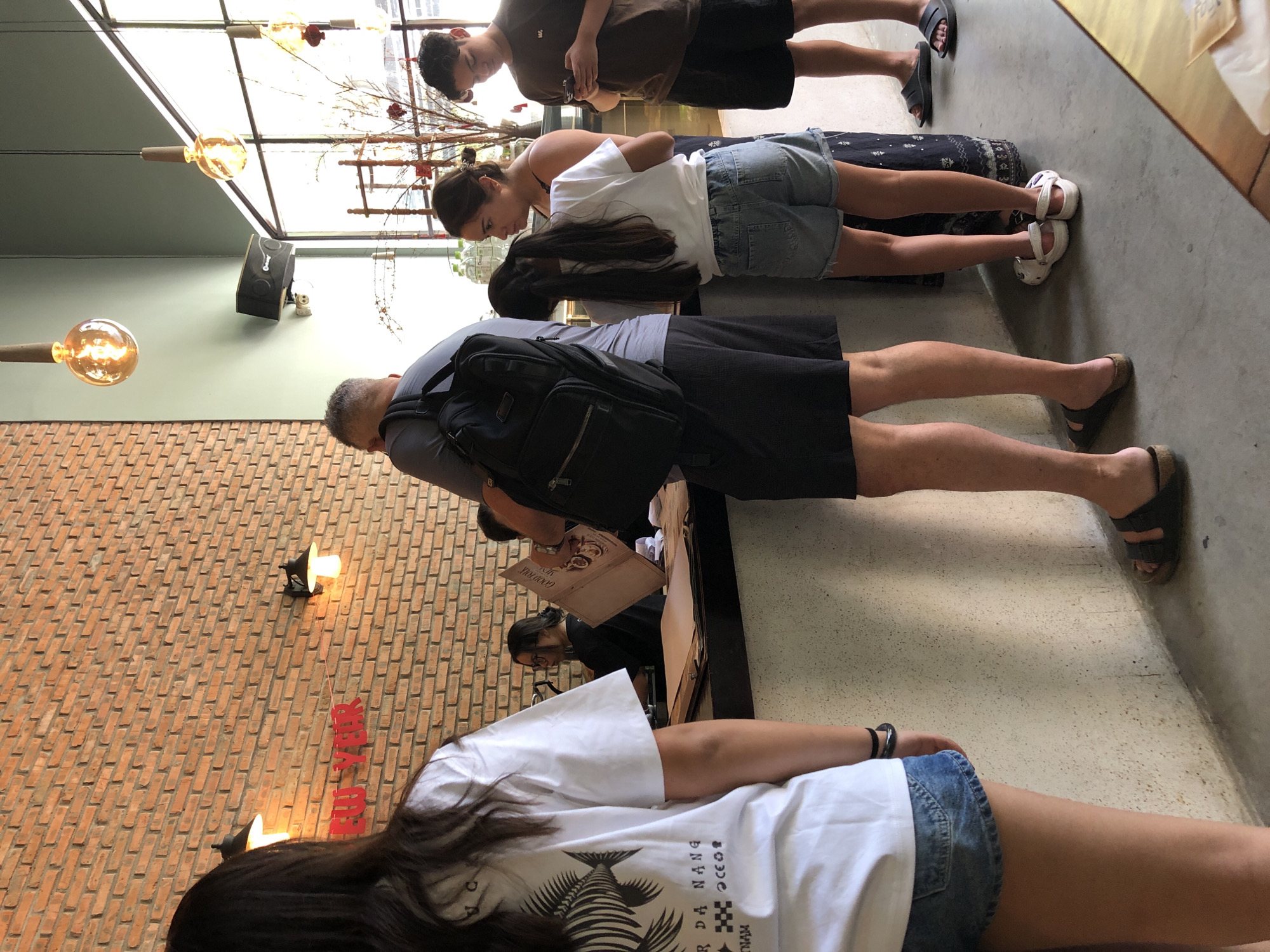 In Đà Nẵng, Vietnam, a group of people stand in line at a counter, likely in a cafe or restaurant. The counter is made of a speckled material, and on it are papers, a menu, and bottled water. Behind the counter, a woman with glasses is visible. The people in line include a man with a backpack, a woman with dark hair, a young girl in denim shorts, and a young man on his phone. The environment features exposed brick walls and modern pendant lighting.