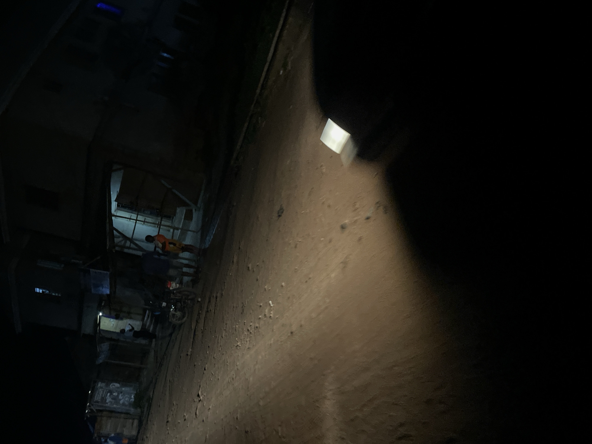 The image depicts a nighttime scene on a dirt road in Lokoja, Nigeria. In the background, several buildings are visible, appearing to be small shops or residences with corrugated metal roofs. One building has an open doorway with light emanating from within. Several people are present, with some gathered near a motorcycle parked on the side of the road. A bright light, possibly from a vehicle's headlight, illuminates a portion of the road in the foreground, casting shadows and creating a stark contrast with the dark surroundings. The ground is uneven and dusty.