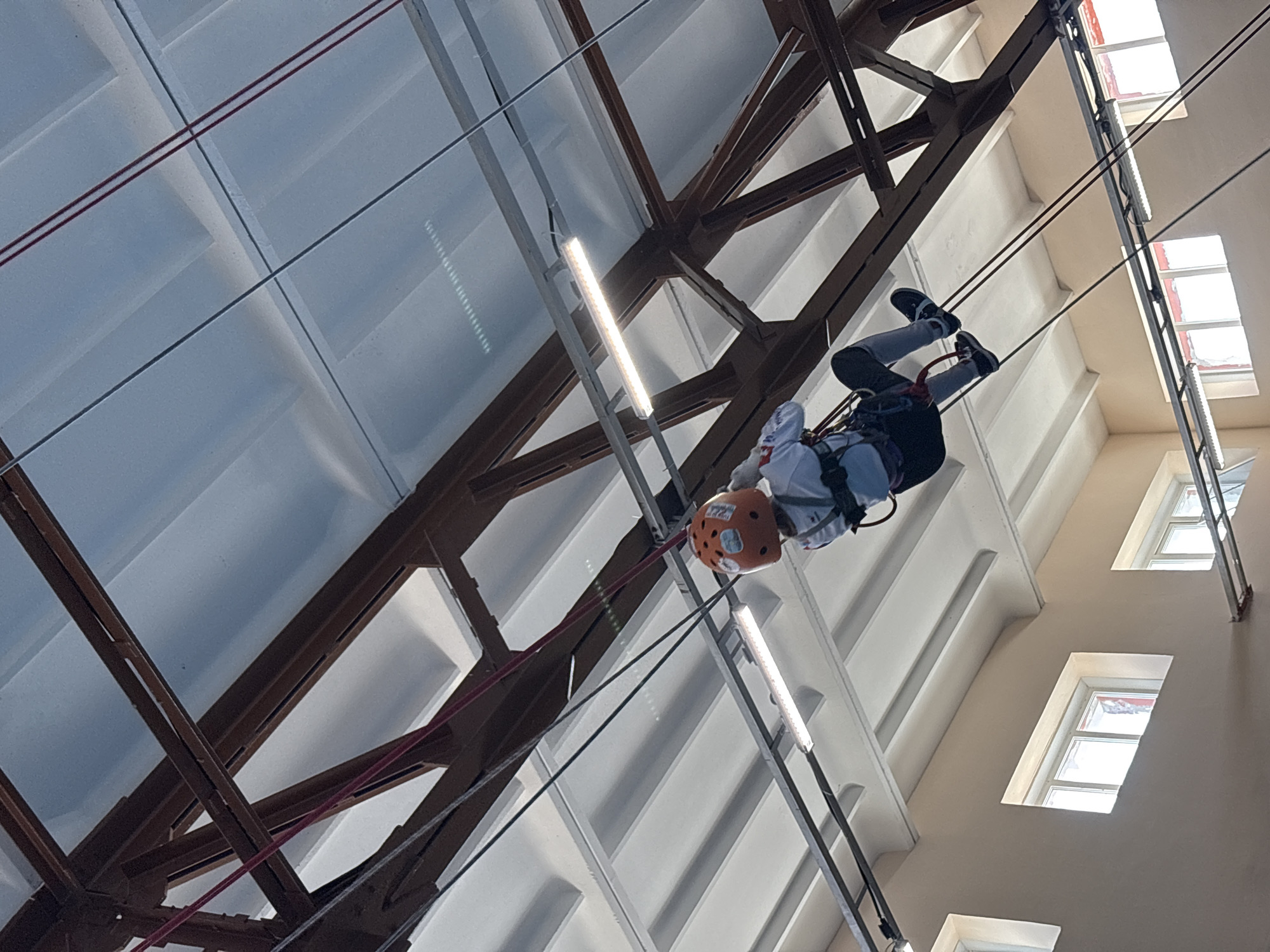 A person wearing an orange helmet is suspended upside down from ropes in what appears to be an indoor space with a high ceiling. The person is wearing grey pants, dark shoes, and a white shirt. The suspension system is attached to a structural metal framework with beams and wires overhead. The ceiling is white with integrated lighting fixtures. Along one side of the image are windows set into a beige-colored wall. The scene appears to be inside a building, potentially a gymnasium or training facility. While it is not clear to pinpoint the specific location, the image is associated with Berezniki, Russia.
