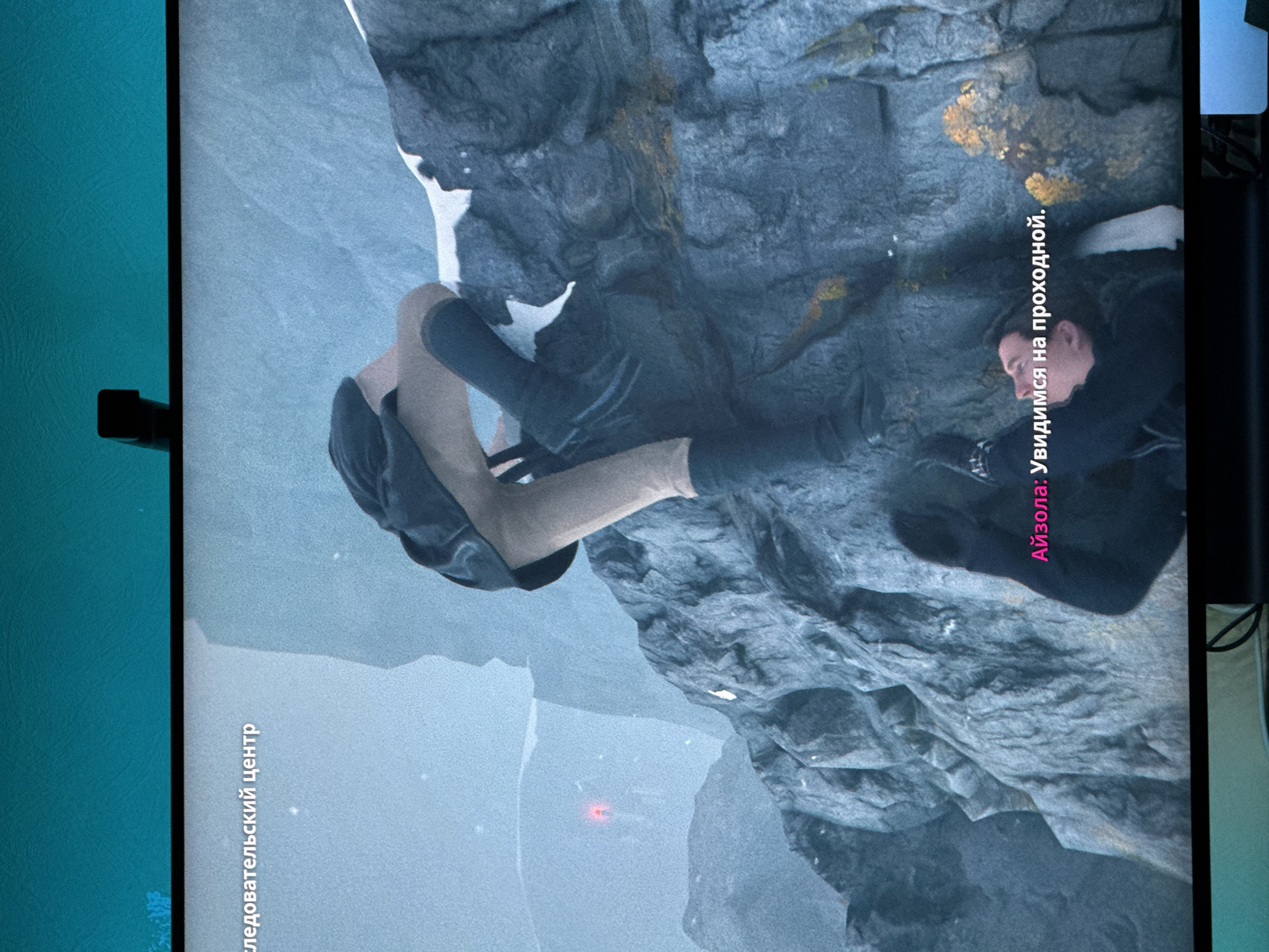 This media file depicts a scene from what appears to be a video game or a cinematic production, set in a rugged, icy mountainous environment. The primary subject is a person, viewed from the waist down and then upwards to their head, dressed in dark clothing and light-colored pants. They are positioned in a precarious, almost horizontal orientation, suggesting they are climbing, falling, or suspended.
The background is dominated by a large, textured rock face, likely a cliff, covered in patches of snow and lichen in various shades of yellow and gray. The ground beneath appears to be snow or ice, with hints of blueish light suggesting a cold, possibly frozen landscape. A faint red light is visible in the lower middle section of the image, its source and purpose unclear within this context.
Two lines of text are visible. The first, oriented vertically on the left side of the image and written in Cyrillic, reads "следовательский центр" which translates to "investigative center". The second line of text, also in Cyrillic and oriented vertically on the right side of the image, reads "Айзола: Увидимся на проходной." This translates to "Aizola: See you at the entrance/turnstile." The text implies a narrative or dialogue component to the scene.
The overall mood is tense and dramatic, evoked by the person's precarious position and the harsh, unforgiving environment. The lighting suggests it could be daytime, but the overcast or atmospheric conditions obscure direct sunlight. The location cues are limited to the mountainous, icy terrain, but the text "investigative center" might hint at a narrative involving an investigation or a research facility within or near this location. The presence of what could be an "entrance/turnstile" mentioned in the dialogue further suggests a structured or perhaps guarded area within this wilderness.