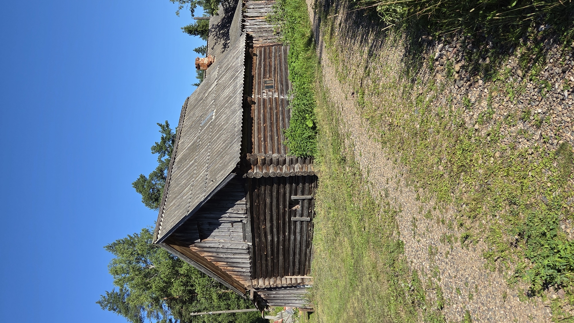 A log cabin with a corrugated metal roof stands under a clear blue sky in Razdol'e, Russia. The cabin is dark brown, constructed from horizontal logs. A small, square window with no glass is visible on the side. Green grass and weeds grow around the base of the structure and on a gravel path leading towards it. Trees, primarily pines, are visible in the background, and a red brick chimney protrudes from the roof of a smaller building further down. The scene appears to be in a rural or semi-rural setting.