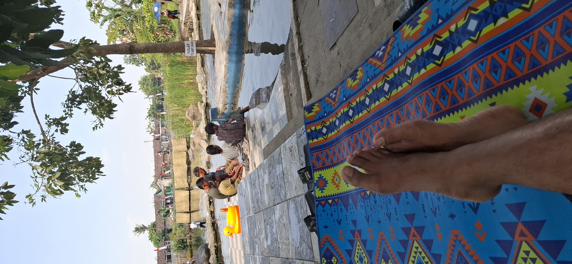 In Wunut, Indonesia, on a sunny day, a group of five individuals, consisting of three adults and two children, are gathered by a shallow, tiled pool. The adults are seated on the tiled edge of the pool, with two of them appearing to converse. One child is sitting with their legs in the water, while the other sits with their back to the camera. A bright yellow duck-shaped float lies on the tiled ground next to them. In the foreground, a person's bare feet rest on a colorful patterned mat. The background reveals a village with houses and Indonesian flags, along with lush greenery and trees. A sign on a tree trunk near the pool reads "KOLAM TERAPIKAN IKAN," indicating a fish therapy pond. The environment suggests a recreational area.