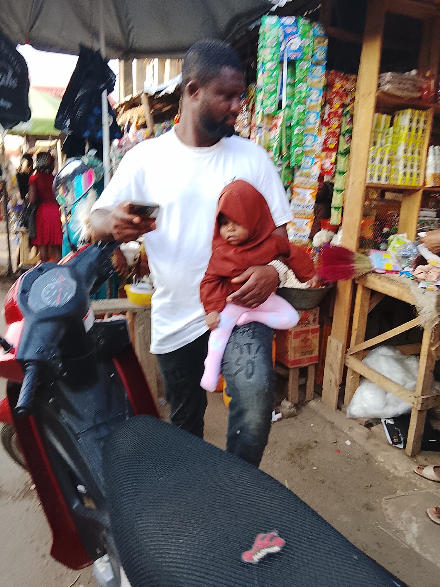 In a bustling outdoor market setting in Akwanga, Nigeria, a man wearing a white T-shirt and black pants holds a small child dressed in a brown hooded jacket and light pink leggings. The child is positioned on what appears to be the man's knee or leg. The man is looking down at a phone he holds in his hand. To the left, a red and black motorcycle is visible. In the background, there are market stalls displaying goods, including stacked packaged items. The ground is a dirt or concrete surface, with additional items and people scattered throughout the scene.