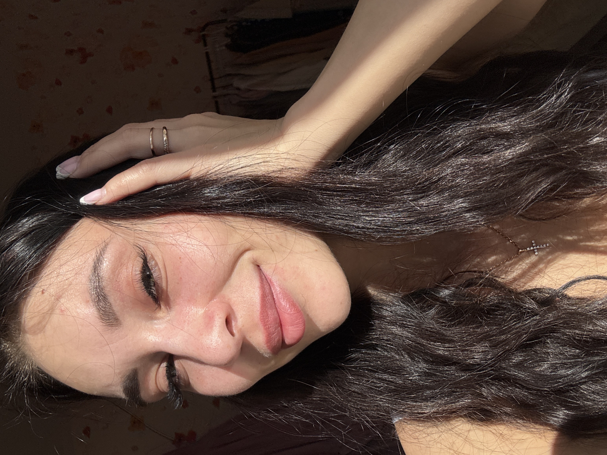 A young woman with dark hair and fair skin is lying down, with her hand resting on her head. She is smiling slightly with her eyes closed. She has two rings on her ring finger, and her nails have a light pink color with white tips. She appears to be wearing a cross necklace. The background is blurred and indistinct, but there's what appears to be a floral patterned surface. The image has warm lighting, creating shadows across her face and arm. There is no information to place this image in Chaikovsky, Russia, or any other specific location.