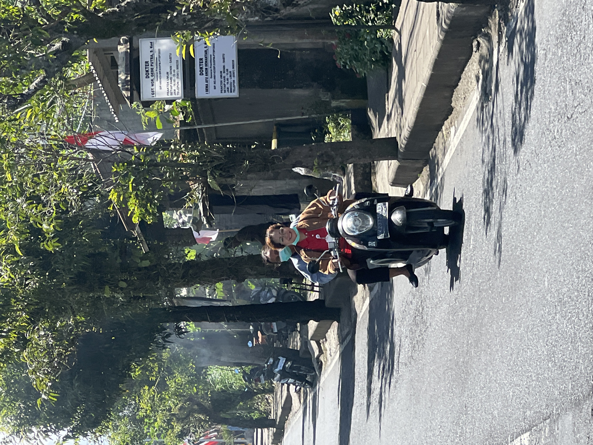 In Payangan, Indonesia, two individuals are riding a motorcycle. The person in front, a woman with dark hair and glasses, wears a red shirt and an orange jacket. She is smiling, looking forward as she operates the motorcycle. Behind her, a second person, whose gender is not clearly discernible due to a blue face mask and dark clothing, sits on the back of the motorcycle. The motorcycle itself is dark-colored with a visible license plate. The scene unfolds on a paved road lined with trees, some bearing the Indonesian flag. On the right side of the road, signs indicate "DOKTER," likely signifying medical practices, with text detailing doctor's names and operating hours. The environment appears to be a sunny day, with shadows cast by the trees on the road. Several other motorcycles are parked on the side of the road in the background.