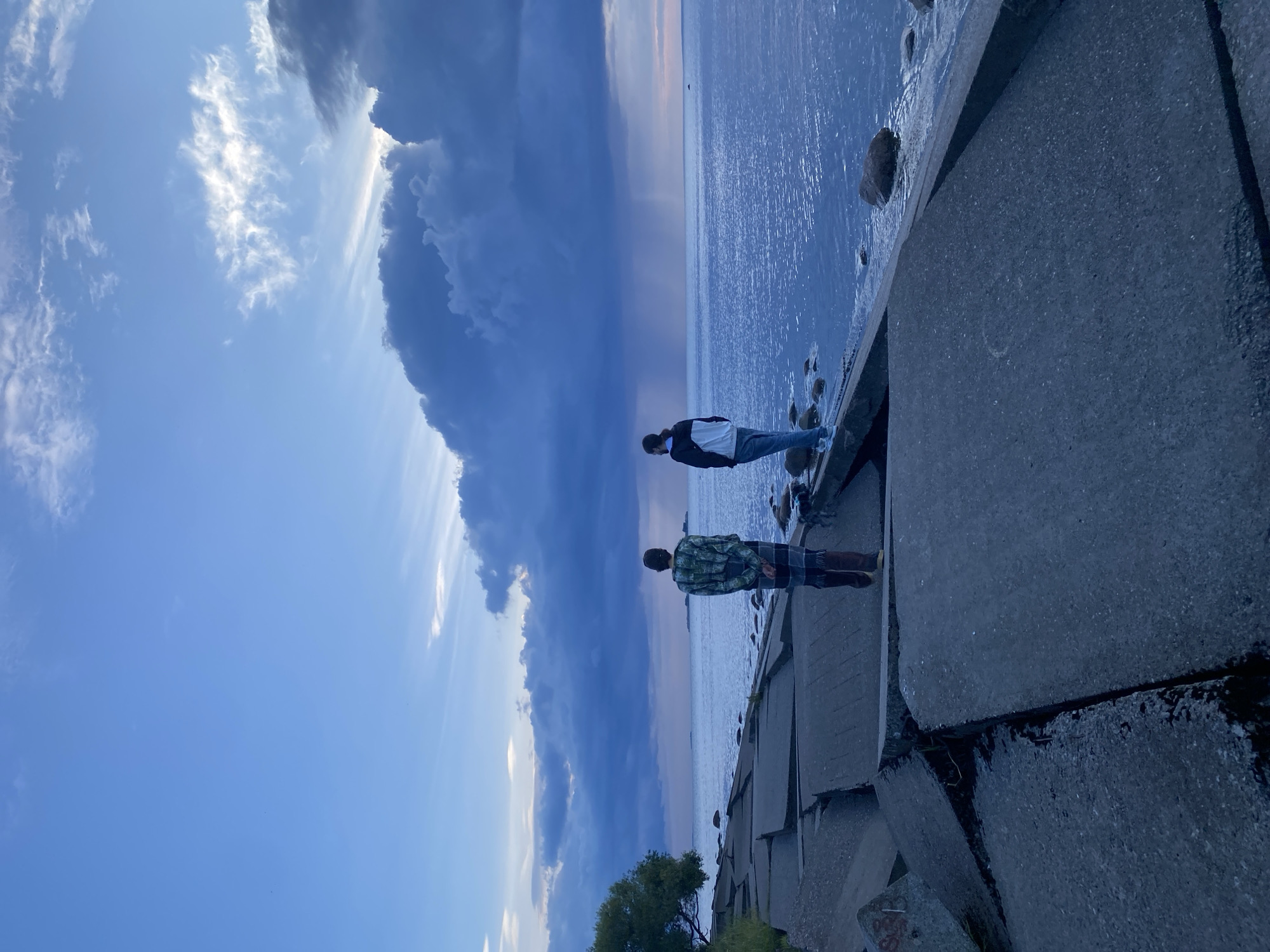 Two individuals are standing on a concrete embankment overlooking a body of water. The embankment is composed of large, irregular concrete slabs, some of which appear broken or dislodged, creating a rough, uneven surface. To the left of the embankment, there is a small tree with green foliage. The water is calm and reflects the sky. The sky above is a gradient of blue, with darker, stormy clouds gathering on the horizon and lighter clouds higher up, with rays of light breaking through. There is a noticeable rain shower occurring in the distance over the water.
The individual on the left is facing away from the camera, wearing a camouflage-patterned jacket and checkered pants. The individual on the right is also facing away from the camera, wearing a dark jacket with white accents and light-colored pants.
This scene is located at the Sestroretsk sea front in Sestroretsk, Russia, characterized by its concrete breakwater and view of the Gulf of Finland. The time of day appears to be late afternoon or early evening, given the lighting conditions.
