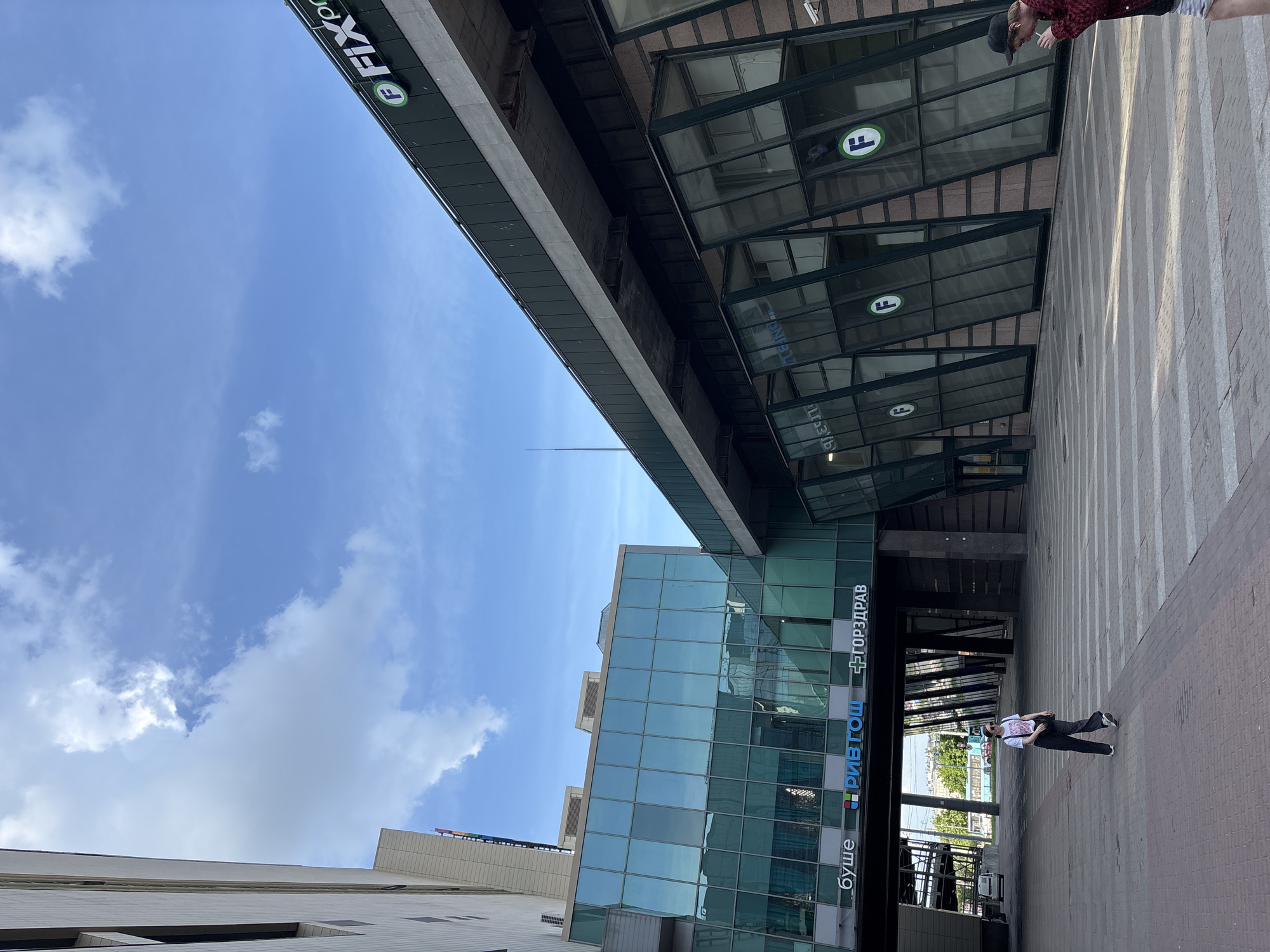 This detailed description is of the Admiralteyskiy Shopping Center at Sadovaya Street 62, Saint Petersburg, Russia.
The image captures an urban exterior scene on a clear, sunny day with scattered white clouds in a bright blue sky. The foreground features a patterned paved area, composed of light and dark grey rectangular pavers, with a section of reddish-brown brick paving leading into a marked pedestrian crossing with white stripes.
Dominating the mid-ground is a modern, multi-story commercial building complex. A distinctive glass-enclosed pedestrian bridge, tinted green-blue, spans over a street, connecting two sections of the building. Visible on the bridge's facade are illuminated signs in Cyrillic for "БУШЕ" (Bushe, a cafe/bakery), "РИВ ГОШ" (Rive Gauche, a cosmetics/perfume store), and "ГОРЗДРАВ" (Gorzdrav, a pharmacy).
To the right, the building exhibits a striking architectural design with multiple dark-framed, outward-sloping glass panels on its lower level, reflecting elements of the environment. Above these panels, a dark green band displays several white-on-green circular "F" logos and the text "FIX price". The upper structure of this building is a dark, possibly concrete or metal, facade with a prominent concrete beam running horizontally along its length.
On the left, a portion of another building with a light-colored, possibly stone or concrete facade featuring horizontal banding is visible. Underneath the pedestrian bridge on the left, a white van is parked. In the background, beyond the bridge and obscured by some trees, a light blue bus is visible on the street. A tall, thin spire-like structure is faintly discernible against the sky in the far background.
Two individuals are present in the scene. A woman is walking in the middle ground, moving from left to right across the paved area. She is wearing a white T-shirt with a graphic design, dark trousers, and sneakers, with sunglasses resting on her head and a dark shoulder bag. On the far right, partially cropped, a man wearing a dark baseball cap, a dark red patterned jacket, and light shorts is holding a white cigarette or vape pen to his mouth.