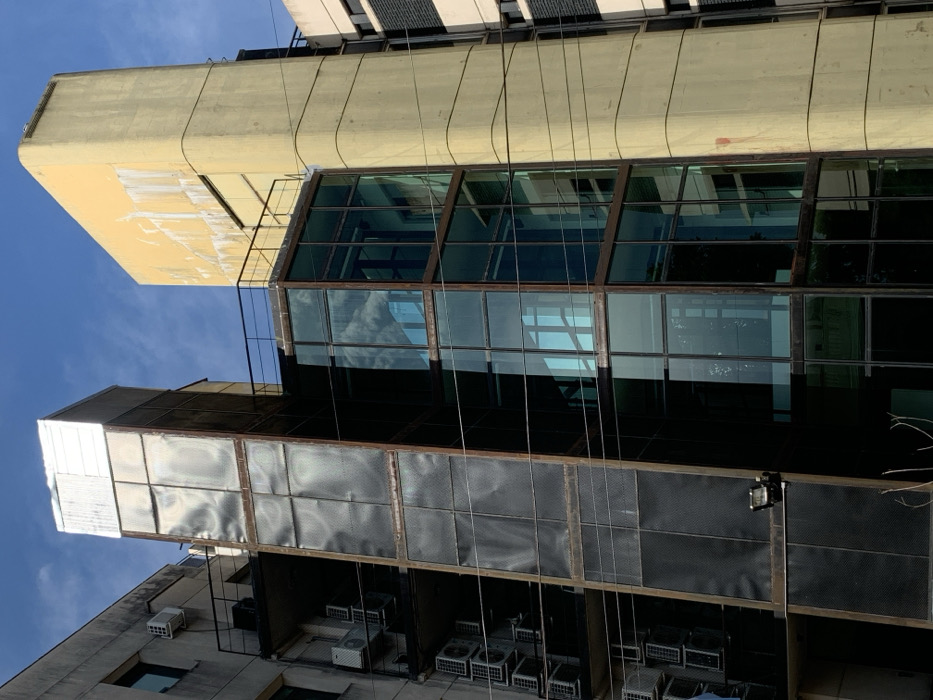 A low-angle shot showcases a building's facade. Its design features a section with large, dark-tinted windows reflecting the sky. Below, another section has a panel-like structure with a metallic frame. Visible beneath this structure are numerous air conditioning units. The sky is a clear blue.