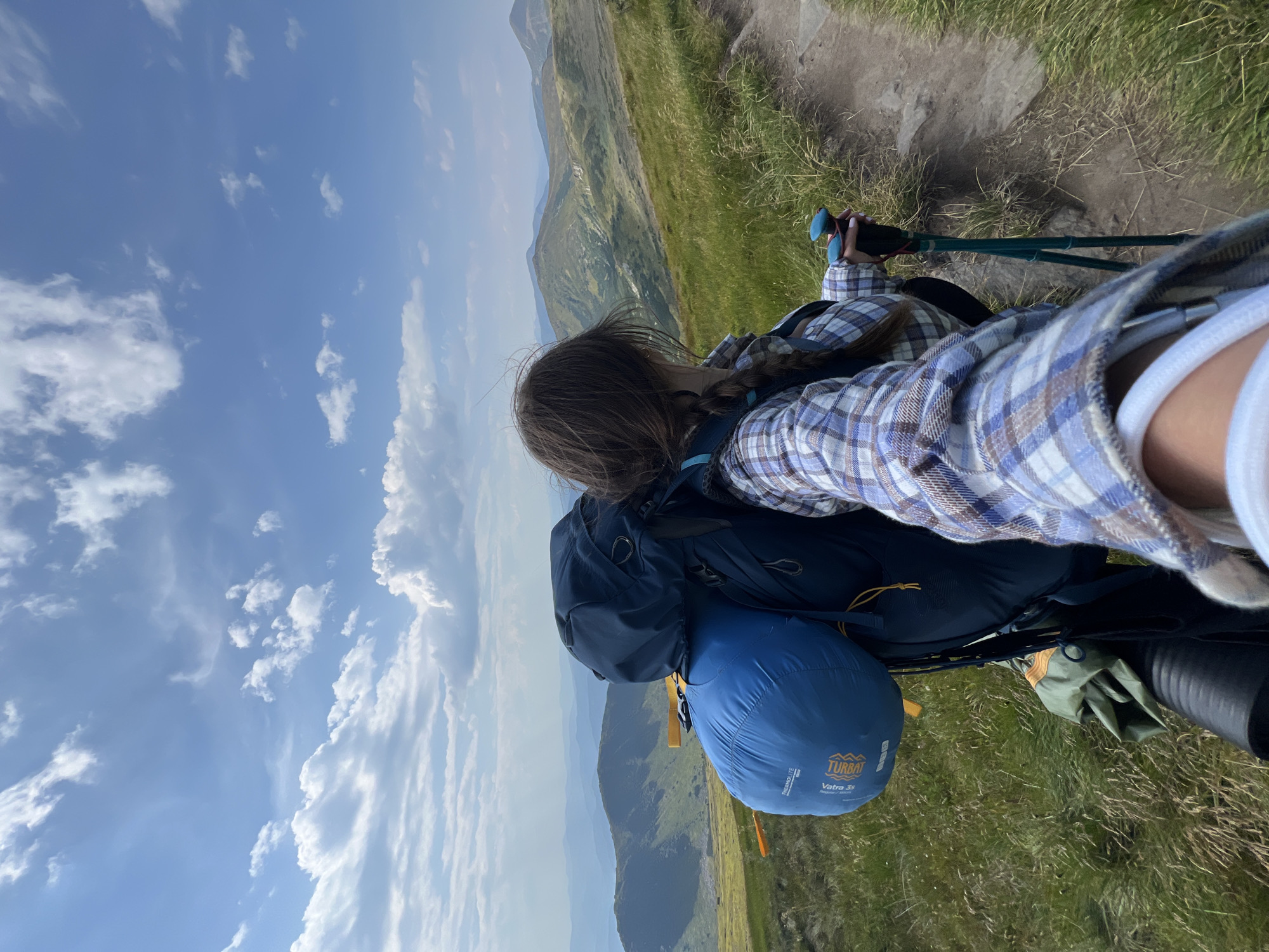 This is a first-person view selfie of a person with a large backpack and trekking poles, standing on a grassy mountain trail. The person is wearing a blue and white plaid shirt and has their hair in a braid. In the background, there are rolling hills and mountains under a blue sky with scattered clouds. The scene is set in the vicinity of Luhy, Ukraine, a village known for its Carpathian mountain landscape. The image captures a moment of hiking or trekking in a natural, mountainous environment.