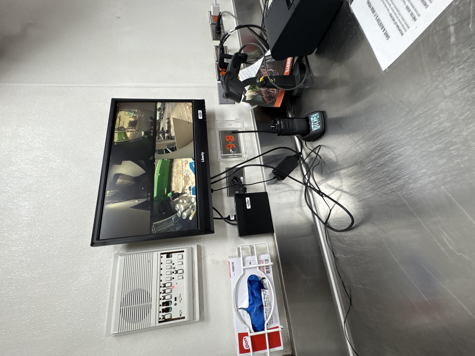 This media file captures a close-up, angled view of a security monitoring station and surrounding equipment, likely in a commercial kitchen or restaurant setting in Panama City Beach, USA. The primary focus is a flat-screen monitor displaying a multi-camera feed, showing various interior views including what appears to be a kitchen area with stacked cans and other equipment. To the left of the monitor is a white intercom or control panel with numerous buttons.
Below the monitor, a box of blue vinyl gloves is mounted in a white wire rack. To the right of the monitor, a silver metal countertop holds several electronic devices. These include a two-way radio with "KITCHEN" clearly visible on its side, a black electronic device, and a charger. Power outlets are visible on the white wall behind the equipment. A paper menu, likely from "DAVE & BUSTER'S," is also present on the countertop, with visible text listing menu items such as "MOUNTAIN OF NACHOS" and "PRETZEL DOOD." The overall scene suggests a functional, operational area, possibly during a business day, given the presence of the intercom and security setup. No people are visible in the frame. The lighting appears to be artificial, typical of an indoor environment.
