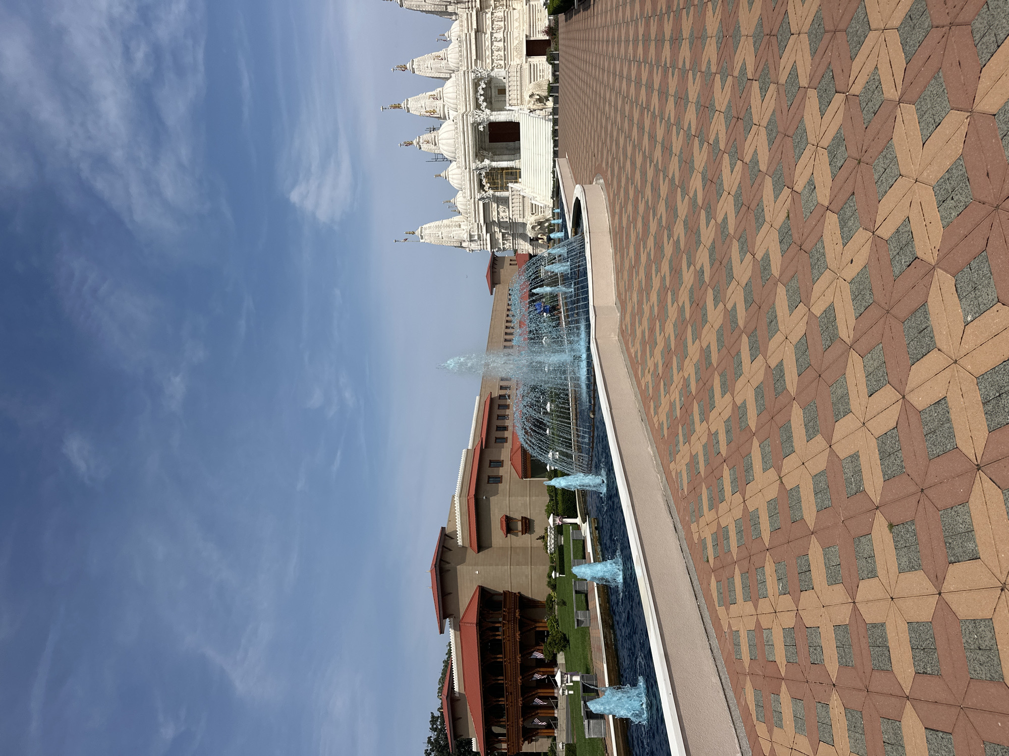 The image depicts the BAPS Shri Swaminarayan Mandir in Bartlett, Illinois, a suburb of Chicago, United States of America. The foreground features a patterned brick walkway with terracotta and brown rectangular bricks interspersed with darker grey square tiles. To the left of the walkway is a long, rectangular fountain with bright blue water spraying upwards from multiple jets, creating a misty effect.
In the middle ground, behind the fountain, is a large, modern building with a light brown facade and a red-tiled roof. This structure has a covered balcony supported by dark wooden beams and features several American flags displayed on poles. Further to the right, and dominating the right side of the image, is the white marble BAPS temple. This ornate structure has multiple domes, intricate carvings, and elaborate spires, with a wide staircase leading up to its entrance. Two white elephant statues flank the base of the stairs.
The background consists of a clear blue sky with wispy white clouds. The scene is bathed in bright daylight, suggesting it was taken on a sunny day. A few people can be seen in the distance, near the entrance of the temple, but they are not distinct enough to identify any specific actions or interactions. The overall impression is one of a well-maintained, architecturally significant religious site.