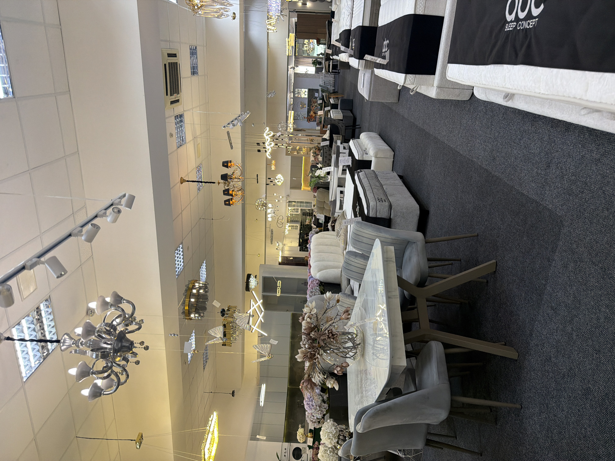The image depicts the interior of a furniture showroom, likely located in Trușeni, Moldova. The showroom displays a variety of furniture, including sofas, beds, mattresses, and dining sets. Several chandeliers and pendant lights hang from the ceiling, illuminating the space. The flooring appears to be dark carpet. In the foreground, a marble-topped dining table with four light gray upholstered chairs is visible. Further into the showroom, more furniture, including several beds with mattresses, is arranged. Decorative floral arrangements are placed on some of the furniture. The overall impression is a well-lit and organized retail environment.
