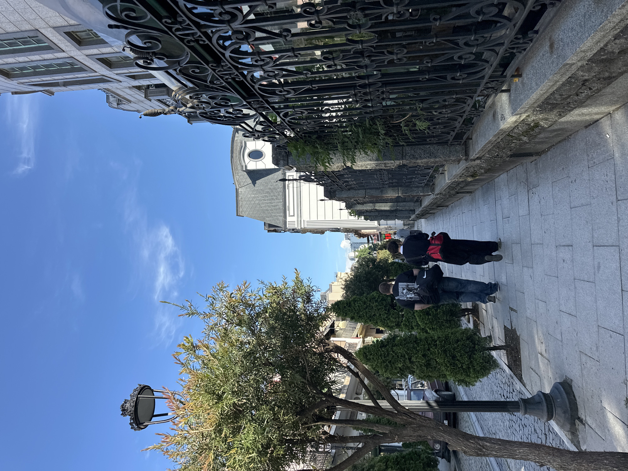 The image depicts a street scene in Batumi, Georgia, characterized by its blend of architectural styles and urban elements. The scene is captured at street level and looks up at the sky. On the right side of the frame, a pedestrian walkway is paved with large, rectangular tiles. Two individuals are walking along this path, their backs facing the viewer. The first person is wearing jeans and a black shirt with a graphic print, carrying a backpack with a Nike logo. The second individual is dressed in black clothing with a red bag.
The left side of the scene is characterized by a tree with a traditional street lamp post at its base. Beyond the tree, buildings can be seen. The sidewalk is lined with decorative wrought iron fences. In the background, a building with a prominent arched window and a gray tiled roof is visible. Other buildings are also visible in the distance, indicating a bustling urban environment. The sky is blue with scattered, wispy clouds.