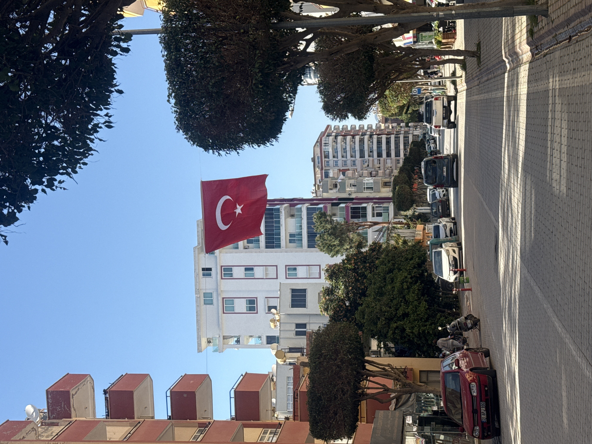 The image captures a street scene, likely in Turkey, given the prominently displayed Turkish flag. The main subject is a large, vibrant red Turkish flag with a white crescent moon and star, hanging vertically from a pole against a clear blue sky.
The scene features multiple multi-story buildings, predominantly white and light-colored, with numerous windows and balconies. Some buildings have modern architectural elements, while others show more traditional designs. On the left side, parts of a building's rooftop with three distinct, reddish-brown cuboid structures are visible.
Lining the street are large, heavily pruned trees, creating a green canopy and casting shadows. The street itself is paved with asphalt, and a patterned brick sidewalk runs alongside it on the right. Bright sunlight is evident, casting strong, elongated shadows across the sidewalk and street, suggesting it's either morning or late afternoon.
Several vehicles are parked along the street. In the foreground on the right, a red sedan with the license plate "31 APJ 990" is clearly visible. Further down the street, a dark hatchback, a white pickup truck, and at least two motorcycles or scooters are parked.
Visible text on a storefront or building sign includes "GLASS BALCONY SYSTEM," accompanied by its Arabic translation "زجاج بلكون" and Russian translation "стеклянный балкон," indicating a business related to balcony installations. The location appears to be an urban or semi-urban area, bustling with parked vehicles but without direct human activity captured in the frame.