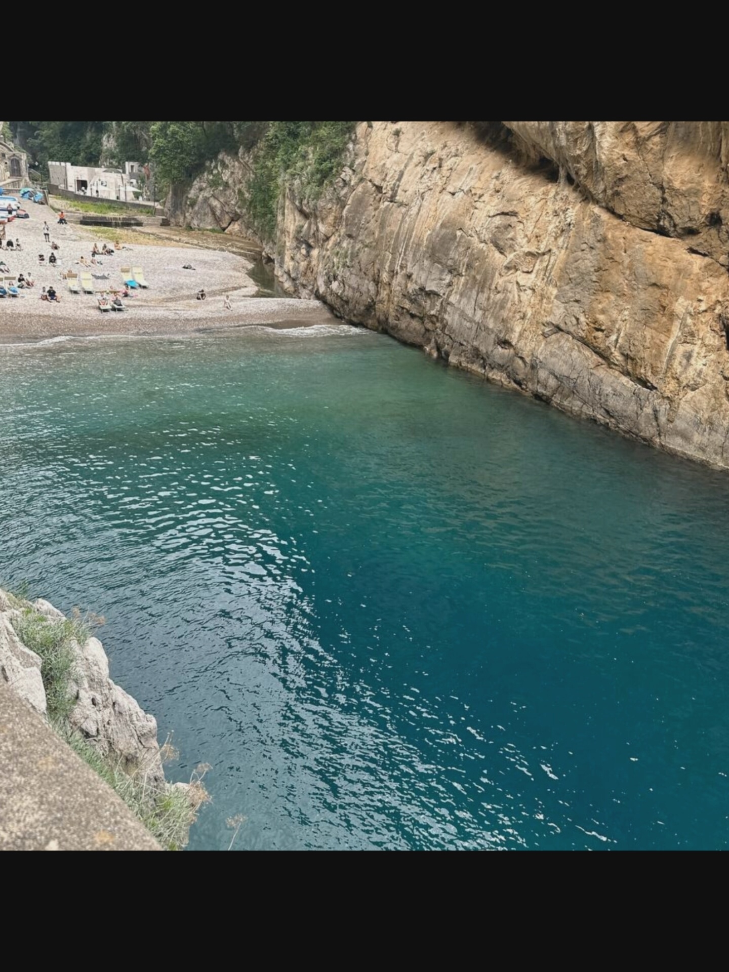 The image depicts a picturesque beach with crystal-clear turquoise water. The beach is a mix of sand and pebbles, with several sunbathers lounging on chairs. A small stream empties into the sea at one end of the beach. The beach is nestled between towering, rugged cliffs, with lush greenery clinging to their sides. In the background, white buildings with red-tiled roofs are visible, suggesting a small coastal village. The overall impression is one of tranquility and natural beauty. It's unlikely this is Norcia, Italy, as Norcia is landlocked and not a coastal town.