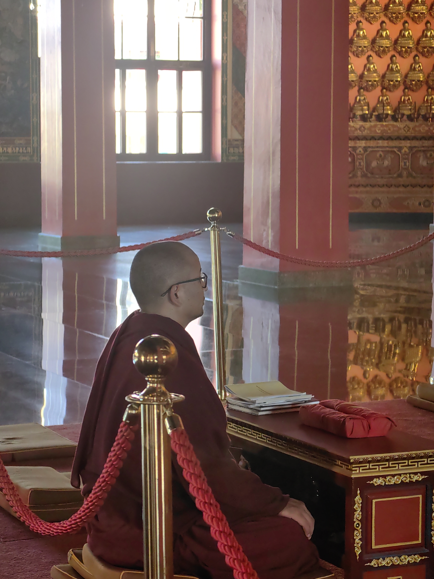 A Buddhist monk, wearing a maroon robe and glasses, sits in profile on a cushion inside a temple. He faces a red wooden desk adorned with gold trim. On the desk are a stack of papers and a red cushion. The monk is surrounded by red rope barriers supported by gold stanchions. In the background, the temple features red pillars, a large window with sunlight streaming through, and a wall decorated with rows of small golden Buddha statues. The floor is reflective, showing the temple's architecture. The location is identified as a temple in Parsa, Nepal.