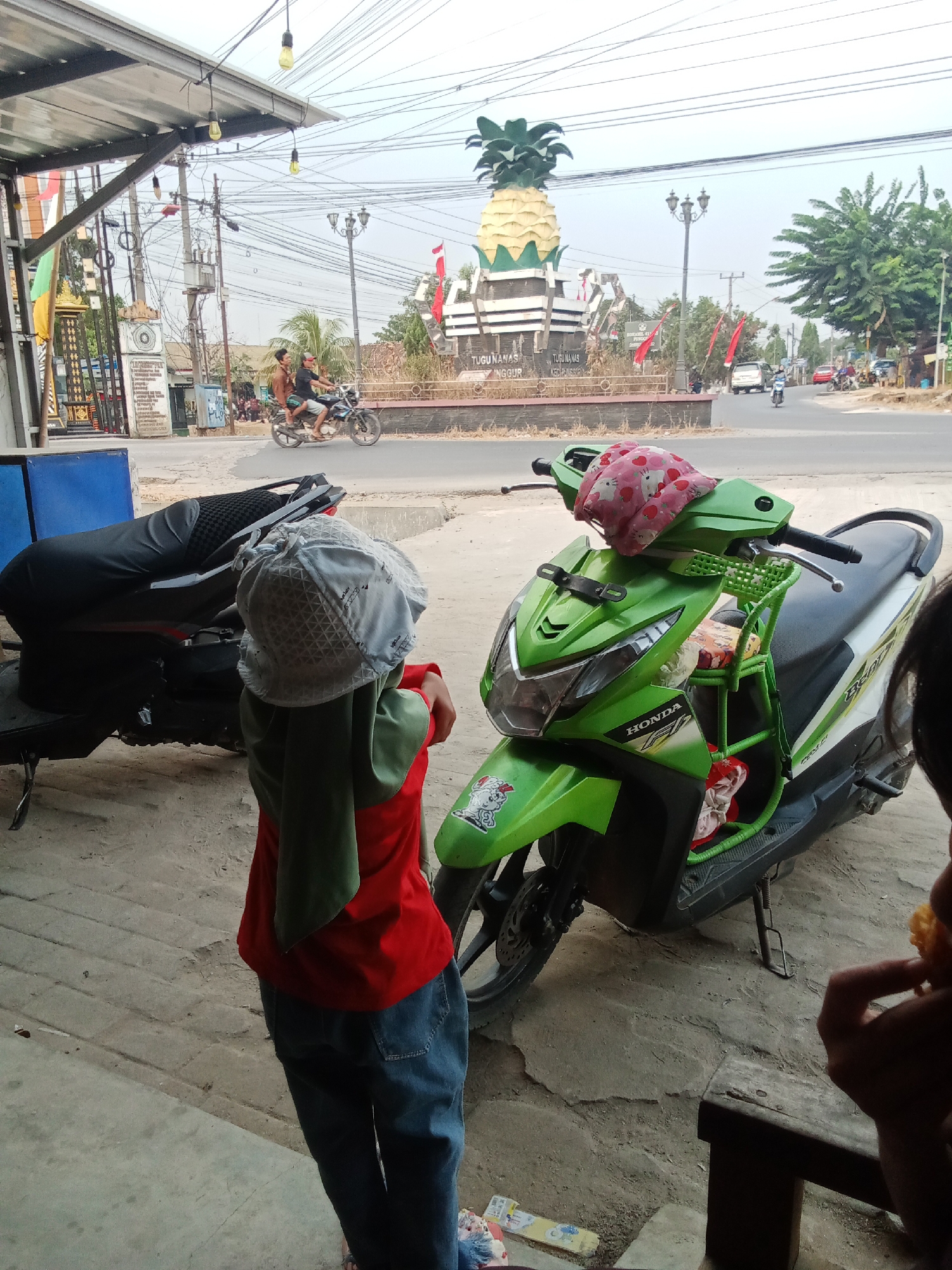In Toto Katon, Indonesia, a child wearing a grey hat, a red shirt, and blue jeans stands by a green and white Honda scooter. The scooter is parked on a sandy lot in front of a covered area. In the background, a large pineapple monument is visible, along with a street and other buildings. Two people ride a motorcycle in the distance.
