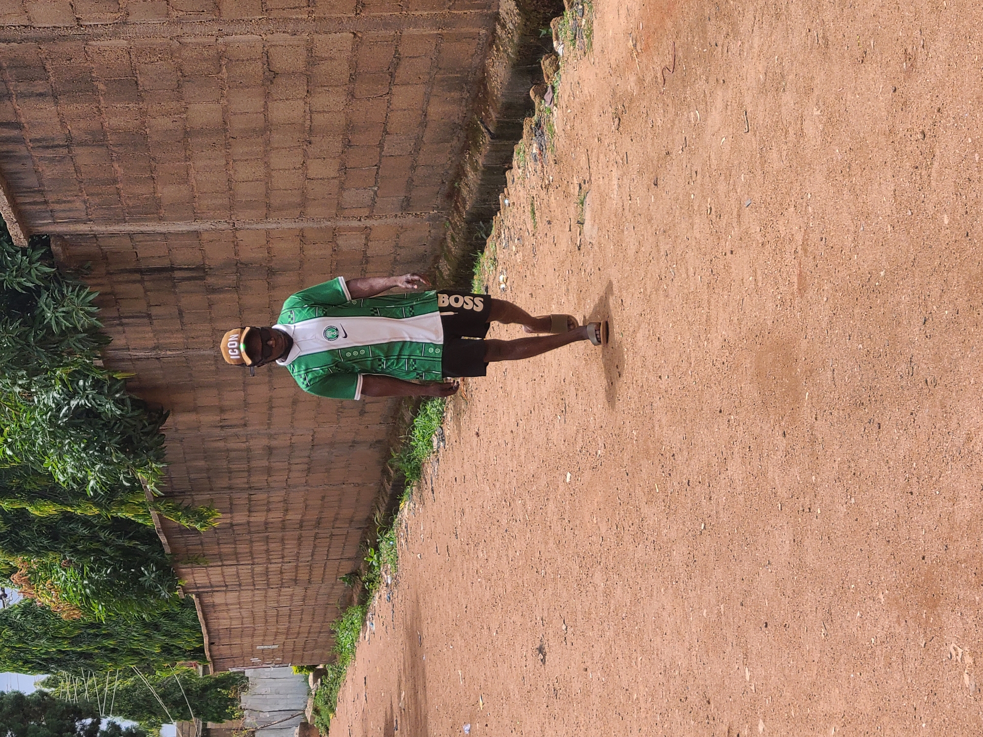 A man stands on a dirt path alongside a brick wall in an outdoor scene. The man wears a green and white soccer jersey, black shorts with "BOSS" written on them, and a brown "ICON" hat. He also wears brown sandals. He is facing the camera, though his body is angled to the right. To the left of the path, there are trees and foliage growing next to the brick wall. The dirt path extends into the distance.
