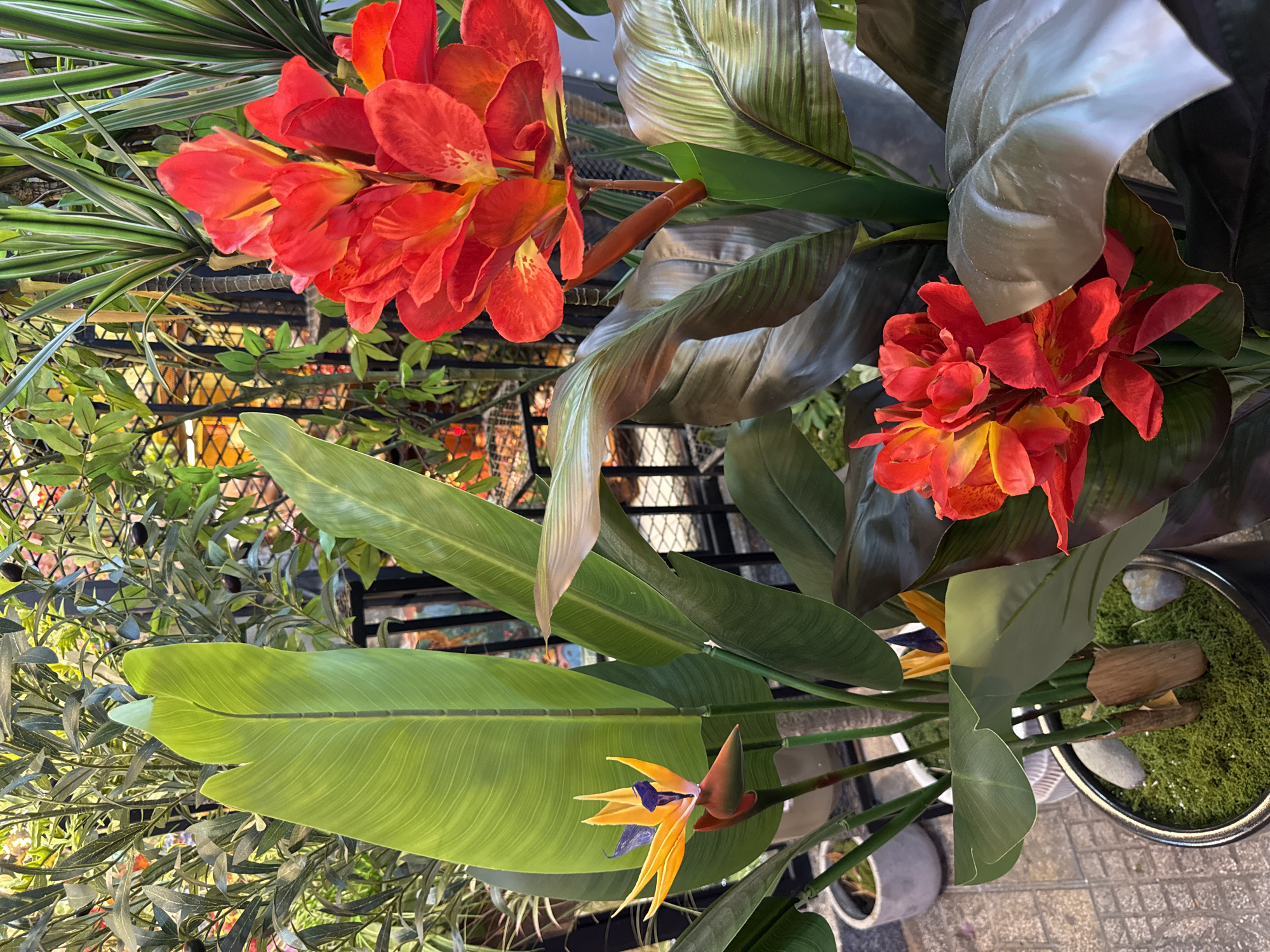 This image shows a close-up of artificial plants, likely displayed outdoors, given the tiled ground and planters visible. In the foreground, a bright orange and yellow bird of paradise flower stands tall with large, green leaves. To its right, a cluster of vibrant red canna lily flowers are in bloom, also surrounded by dark, broad leaves. In the background, various other green plants are visible, including what appears to be an olive tree with black olives, and spiky, striped foliage. A black metal fence is partially visible behind the plants. The overall setting suggests a decorative arrangement, possibly in a garden center, shop, or a well-maintained outdoor space in Nha Trang, Vietnam. There are no people or animals in the image. The lighting indicates it is daytime, with soft shadows suggesting diffused sunlight.